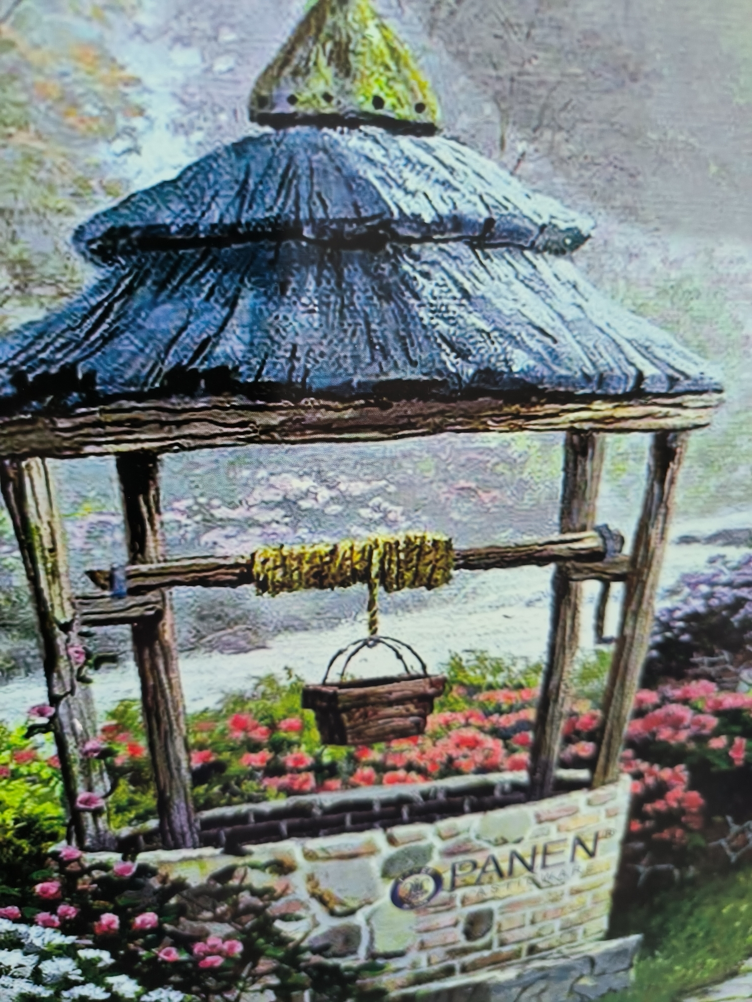 The image depicts a stone well with a wooden roof, a bucket hanging from a rope, and surrounding foliage. The words "PANEN" and "LASTI" are visible on the stone structure of the well. There is no indication of it being in Lhokseumawe, Indonesia. No people, actions, or interactions are depicted. The environment includes flowers and greenery.