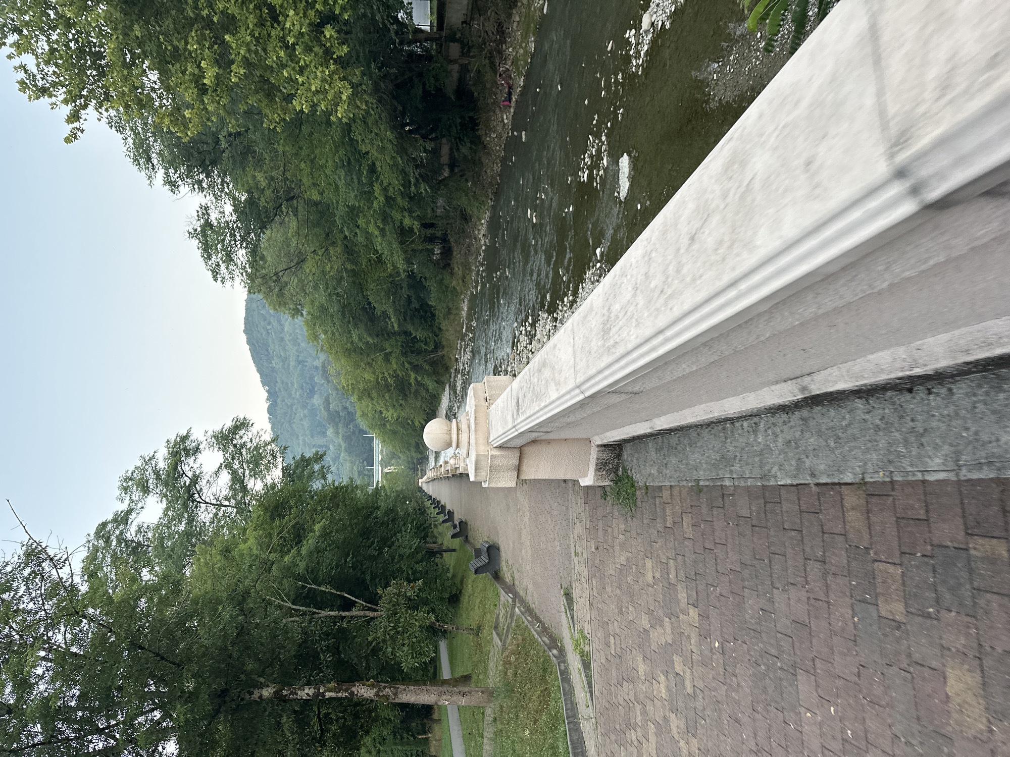 The image shows a paved walkway alongside a river in Sochi, Russia. The walkway is lined with benches and trees. The river is shallow and rocky, with clear water. In the background, a bridge spans the river. The sky is clear and the sun is setting, casting a warm glow over the scene. The overall impression is one of a peaceful and scenic park.