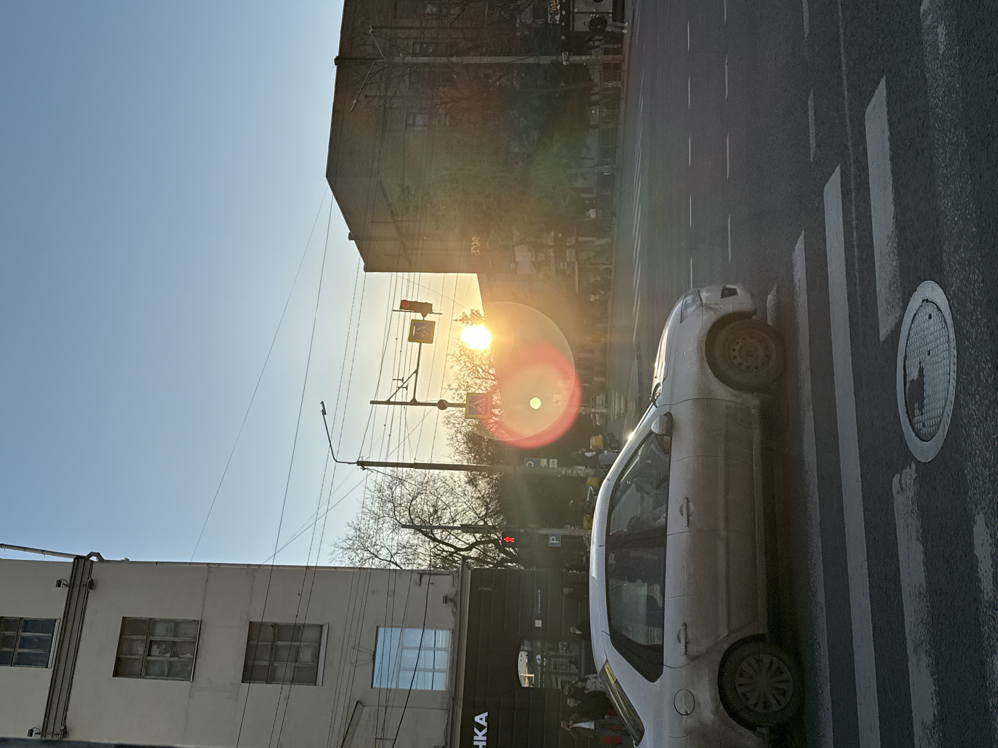 The image shows a street scene in Rostov-on-Don, Russia. A white car is parked on a pedestrian crossing. A manhole cover is visible on the right side of the road. Buildings are present on both sides of the street. A traffic light is illuminated in red. The sun is shining brightly. People are visible in the distance near a building with the Cyrillic word "КА" visible on its facade. Bare trees are silhouetted against the bright sky.