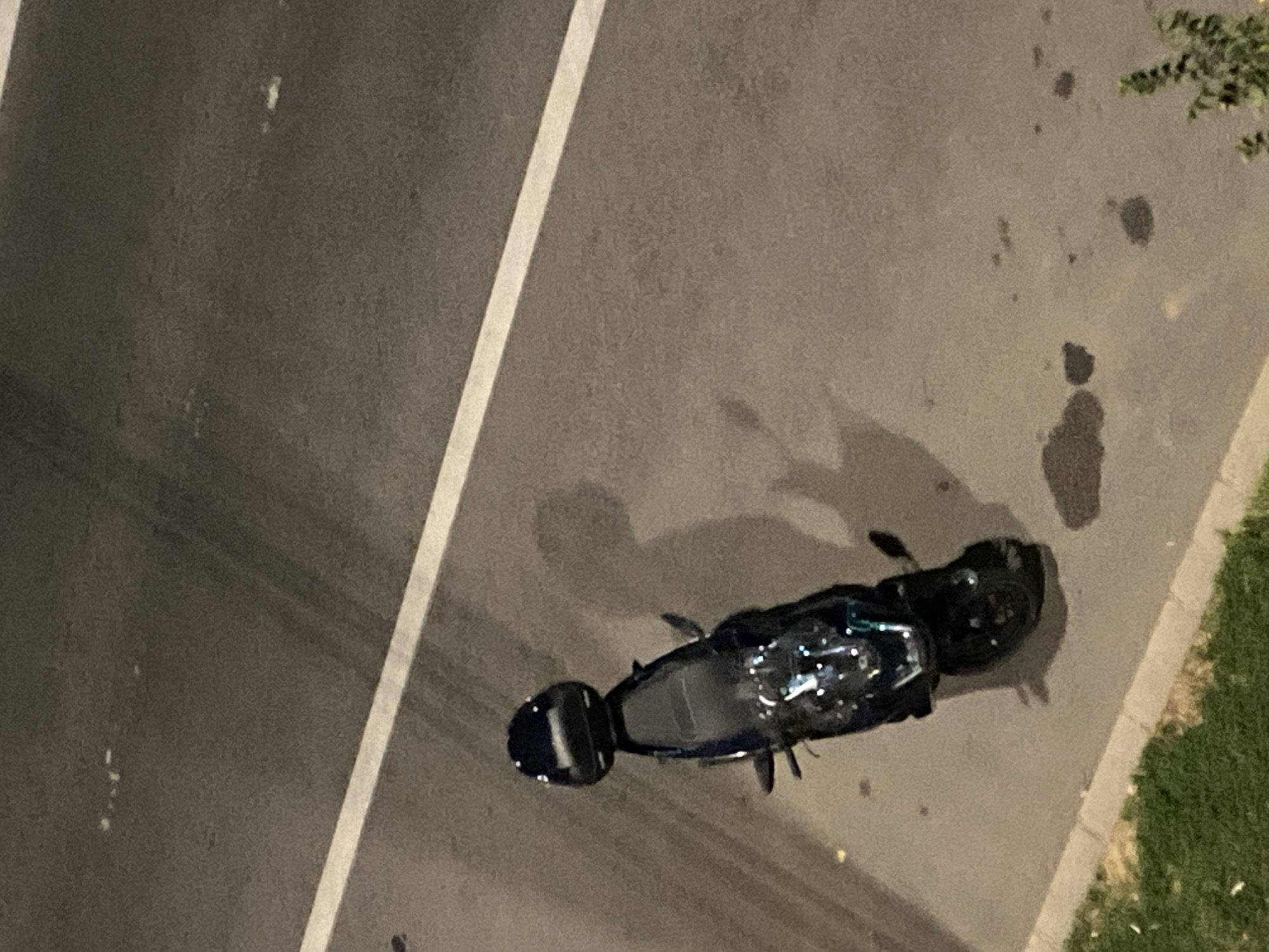 The image, taken from an elevated perspective, shows a black scooter parked on the side of a road in Iași, Romania. The scooter has a black storage box mounted on the rear. A white line, likely a road marking, runs diagonally across the asphalt. The road surface appears to be worn and has some dark patches, possibly oil stains or wet areas. To the left of the road, a strip of grass and a concrete curb are visible. The scene is illuminated by artificial light, casting shadows. The overall impression is of an urban environment at night or dusk.