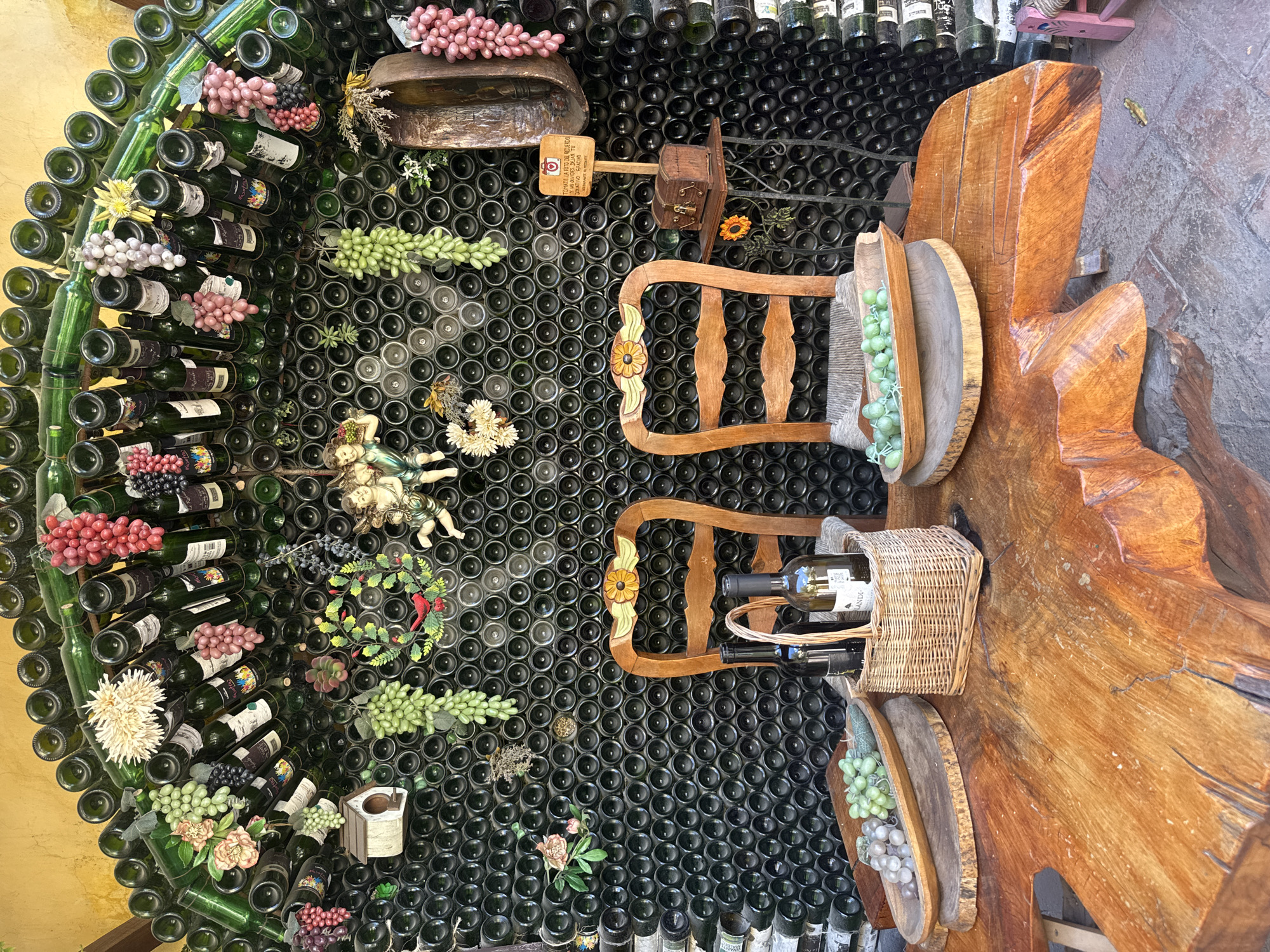 The image shows a decorative wall made of stacked empty wine bottles, likely in Bernal, Mexico. The wall is adorned with artificial grape bunches, flowers, and a small statue of a reclining cherub. In front of the wall is a small seating area with two wooden chairs. One chair contains a bottle of wine in a woven basket. Next to the chairs is a rough-hewn wooden table.