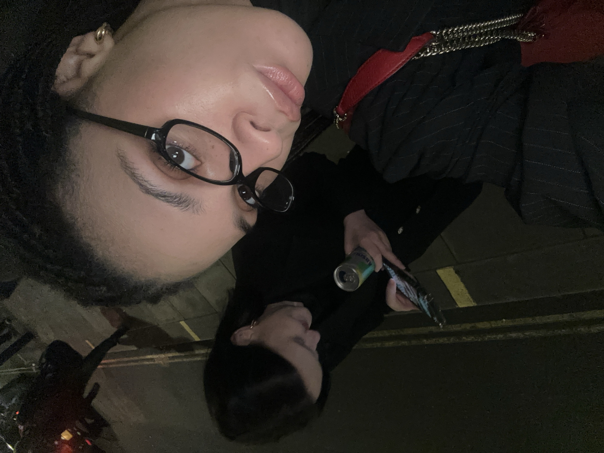 A dark, low-angle street photograph depicts two people at night. The primary subject, in the upper portion of the frame, wears black framed glasses, a gold hoop earring, a dark pinstripe blazer, and a red purse strap is visible. She looks directly at the camera. Below her is a second person, holding a beverage can and a mobile phone, looking in the direction of the first person. The street surface is visible in the lower portion of the photograph, with yellow painted lines. Indistinct objects and figures are in the background. The location appears to be an urban street at night, most likely London, United Kingdom.