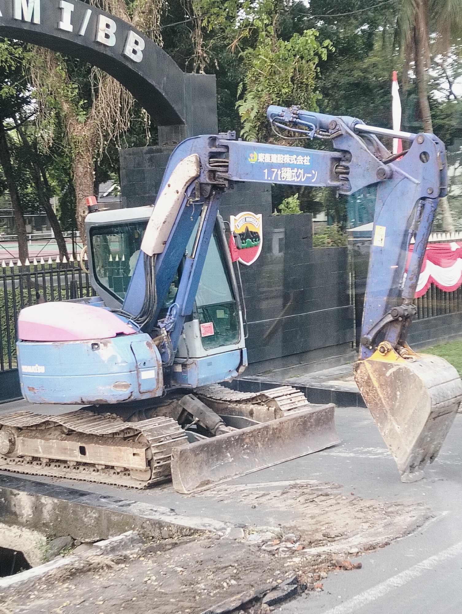 An excavator is parked on a street in front of a building with an archway. The excavator is blue with some pink on its hood. It has a large bucket in front and tracks instead of wheels. There is some debris on the ground around the excavator. Behind the excavator, there is a fence and some trees. On the building, there is a crest with a red and white flag. The archway has the letters "M I / B B" on it. The image appears to be taken during the day. The location is City of Medan, Indonesia.