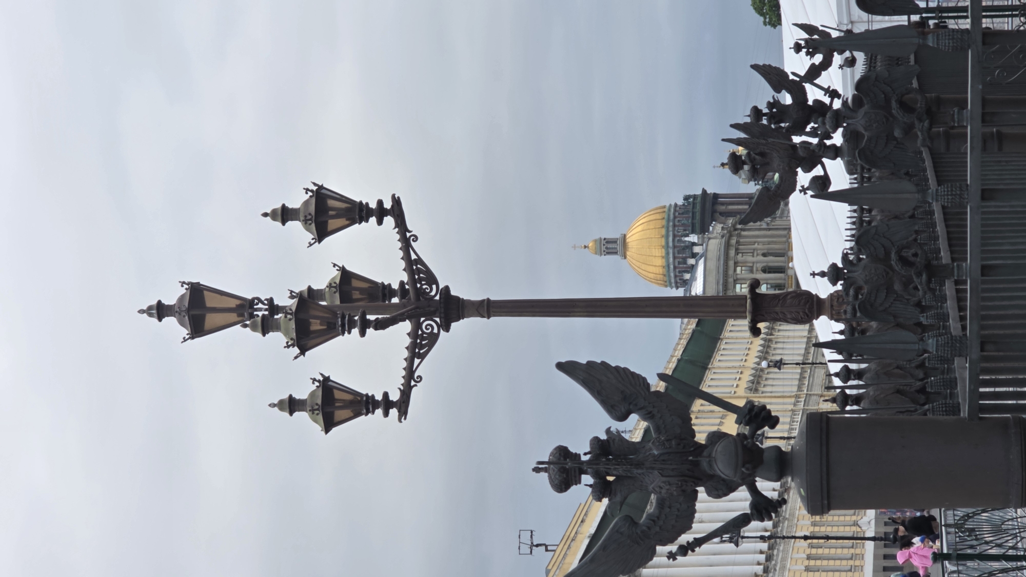 Here is a description of the image:
The image is a view of St. Petersburg, Russia, featuring decorative elements of the city and a glimpse of St. Isaac's Cathedral. A black, ornate lamppost stands prominently in the frame, with multiple lanterns extending from it. To the right of the lamppost is a metal fence adorned with eagles, likely part of a bridge or architectural feature. In the background, the golden dome of St. Isaac's Cathedral is visible, along with sections of buildings with classical architectural details, including columns and ornate facades. The sky above is overcast and gray.