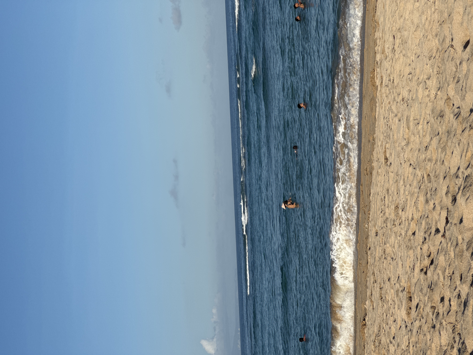 The image depicts a beach scene in Kuta Selatan, Indonesia. The sky is clear and blue. The sea is a deep blue with white waves. Several people are swimming in the water. There is a sandy beach on the right side of the image. One person on a board is near the center of the image.
