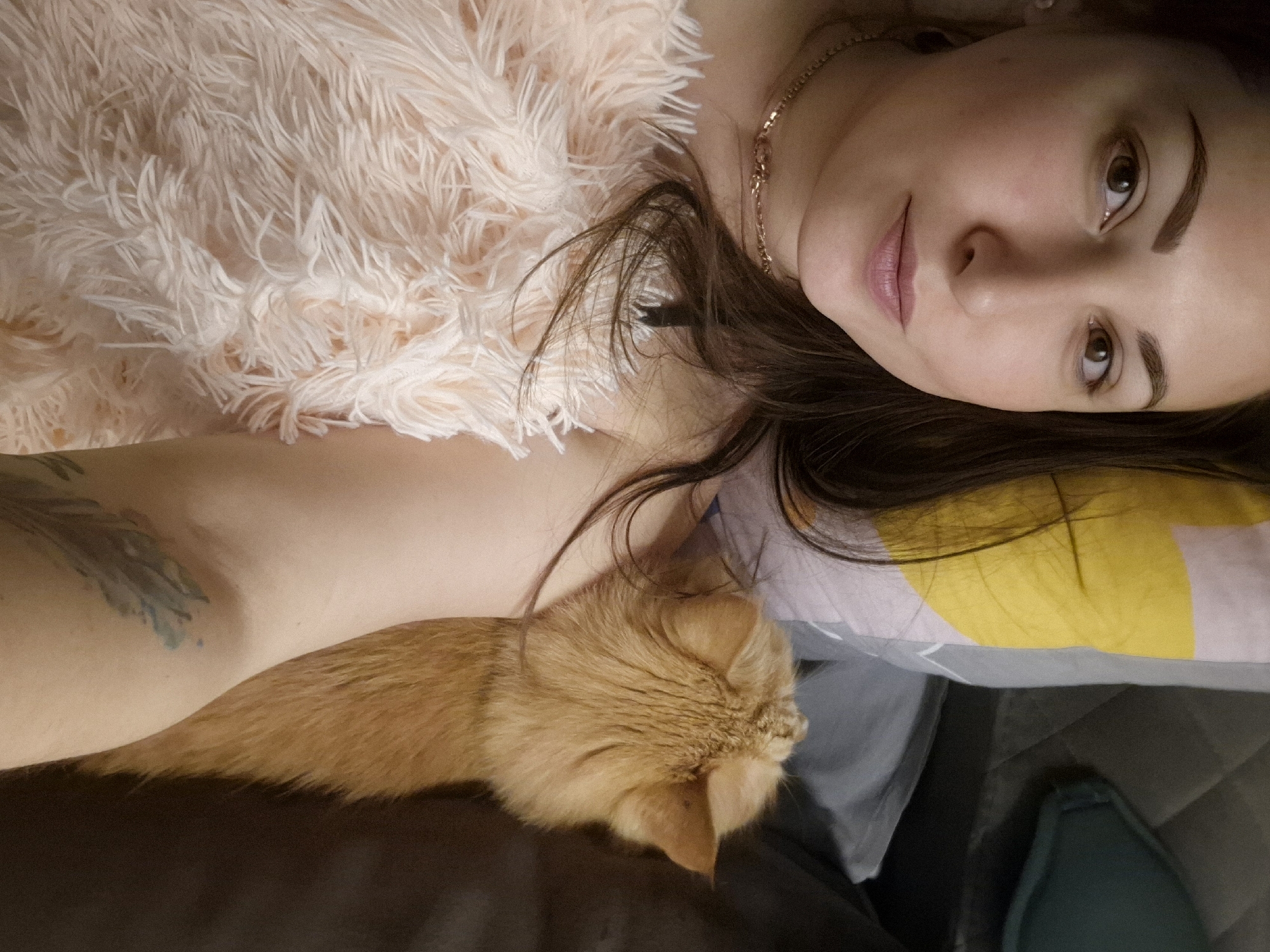 A woman is lying down with an orange cat curled up on her arm. The woman is facing the camera with a slight smile, wearing a fuzzy pink top and a gold necklace. She has brown hair and brown eyes. The cat is facing away from the camera, its fur is a light orange color. The background is a pillow with a yellow and white design, and a dark-colored blanket or sheet. The location is a bed. The city and state are unknown.