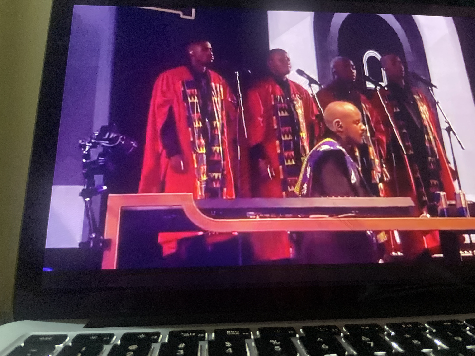 A group of Black men in red robes with patterned stoles stand on a stage, some singing into microphones. A man in the foreground, bald, sits at a keyboard. The stage is lit with purple and white lights. The scene is captured on a laptop screen, with the keyboard visible in the foreground.