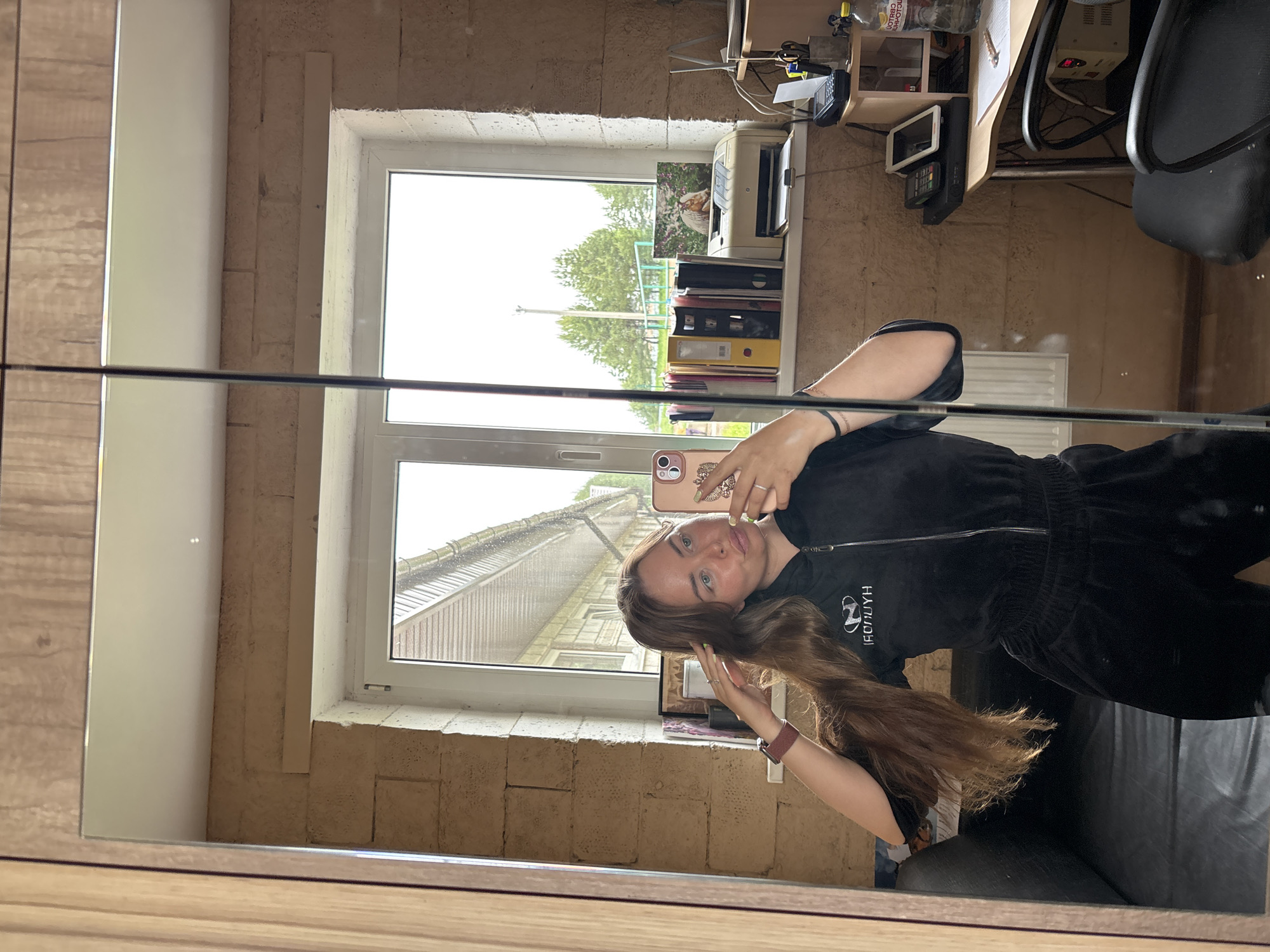 A woman is reflected in a mirror while taking a selfie. She is wearing a dark-colored tracksuit. Her long brown hair is styled to cascade down her front. Behind her, visible through a window, is a view of trees and buildings under an overcast sky. The room has exposed brick walls. A desk cluttered with books and office supplies is partially visible on the right side of the window. The location is Русский Вожой, Russia.