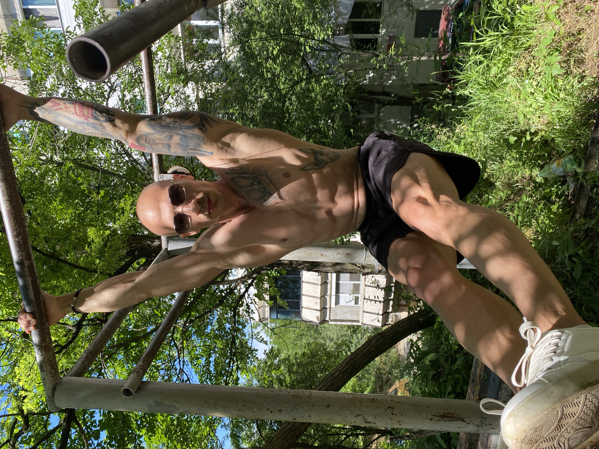 A shirtless, bald man with tattoos on his arms and chest is doing pull-ups on an outdoor horizontal bar. He is wearing sunglasses and black shorts. His white sneakers are visible at the bottom of the frame. The background shows lush green trees and a multi-story building. The scene appears to be in a park or residential area in Pleven, Bulgaria. There is a plastic bottle and some debris on the ground near the base of the pull-up bar.