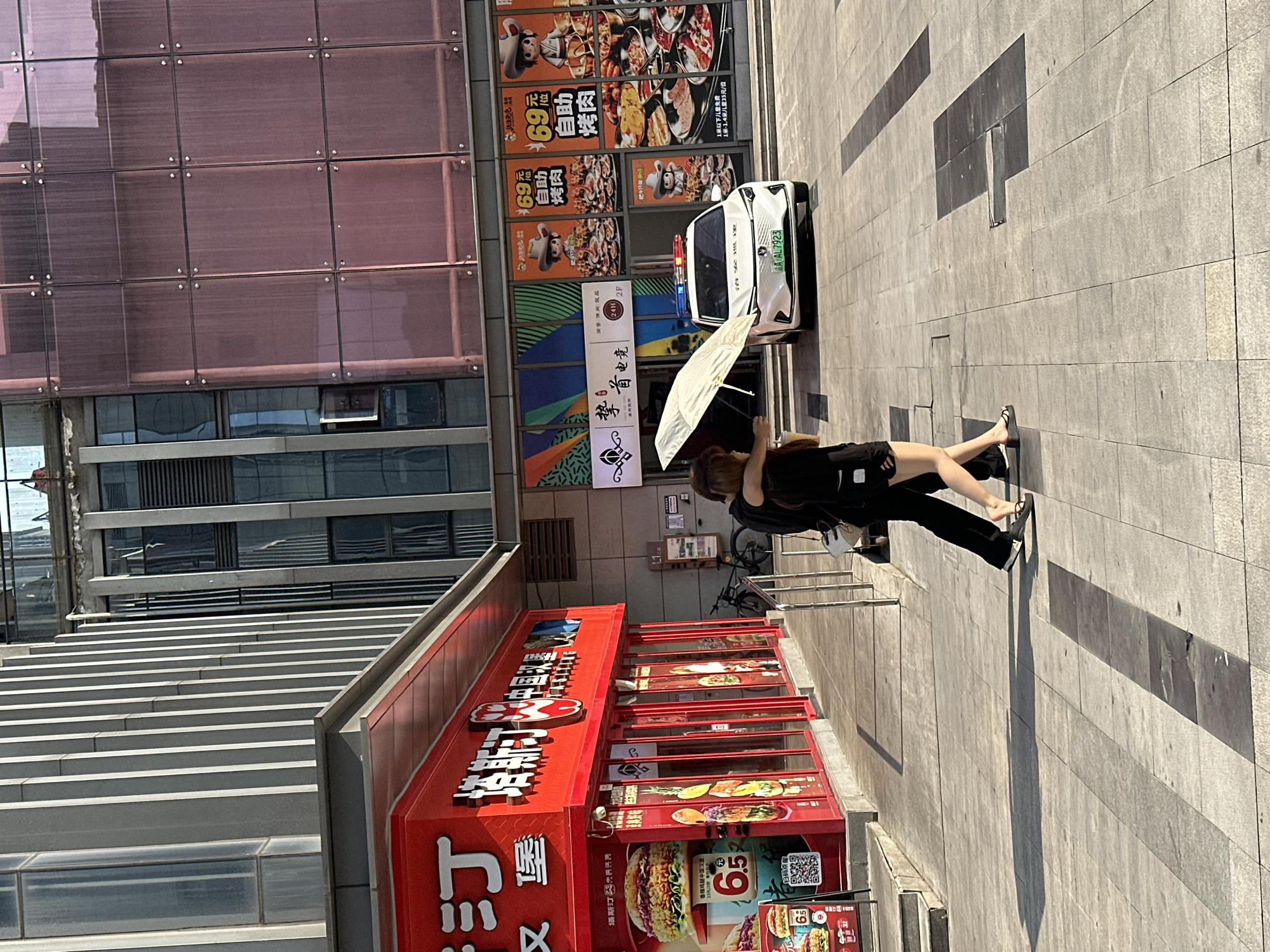 A couple walks under an umbrella on a sunny day in the city of Chongqing High-tech Zone, China. The man has his arm around the woman, and they are walking away from the camera. The woman is holding a white umbrella over them. They are walking on a paved sidewalk in front of a red building with large windows. A white police car with red and blue flashing lights is parked on the street behind them. The police car has a license plate that reads "AL 7923." The background shows modern buildings with glass facades. The ground is paved with large square tiles.