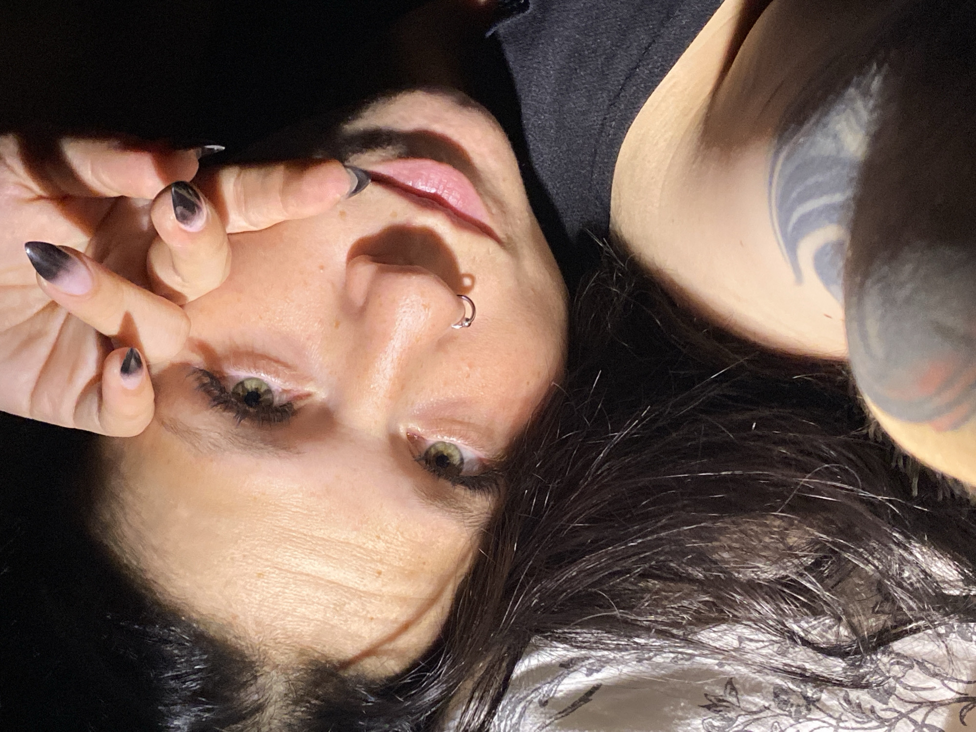 The image is a close-up, overhead, point-of-view shot of a person lying down, looking upwards towards the camera. The person has dark hair, green eyes, and is wearing a black top. Their right hand is raised, with fingers obscuring the upper part of their face. The person has dark, pointed fingernails with a gradient of black and pink polish. A silver nose ring is visible on the left nostril. Sunlight casts dramatic shadows across the person's face. The background is dark, with a hint of a patterned fabric on the left, possibly a pillow or blanket. A tattoo is visible on the person's shoulder. The image does not provide any information about the city of Chaikovsky, Russia.