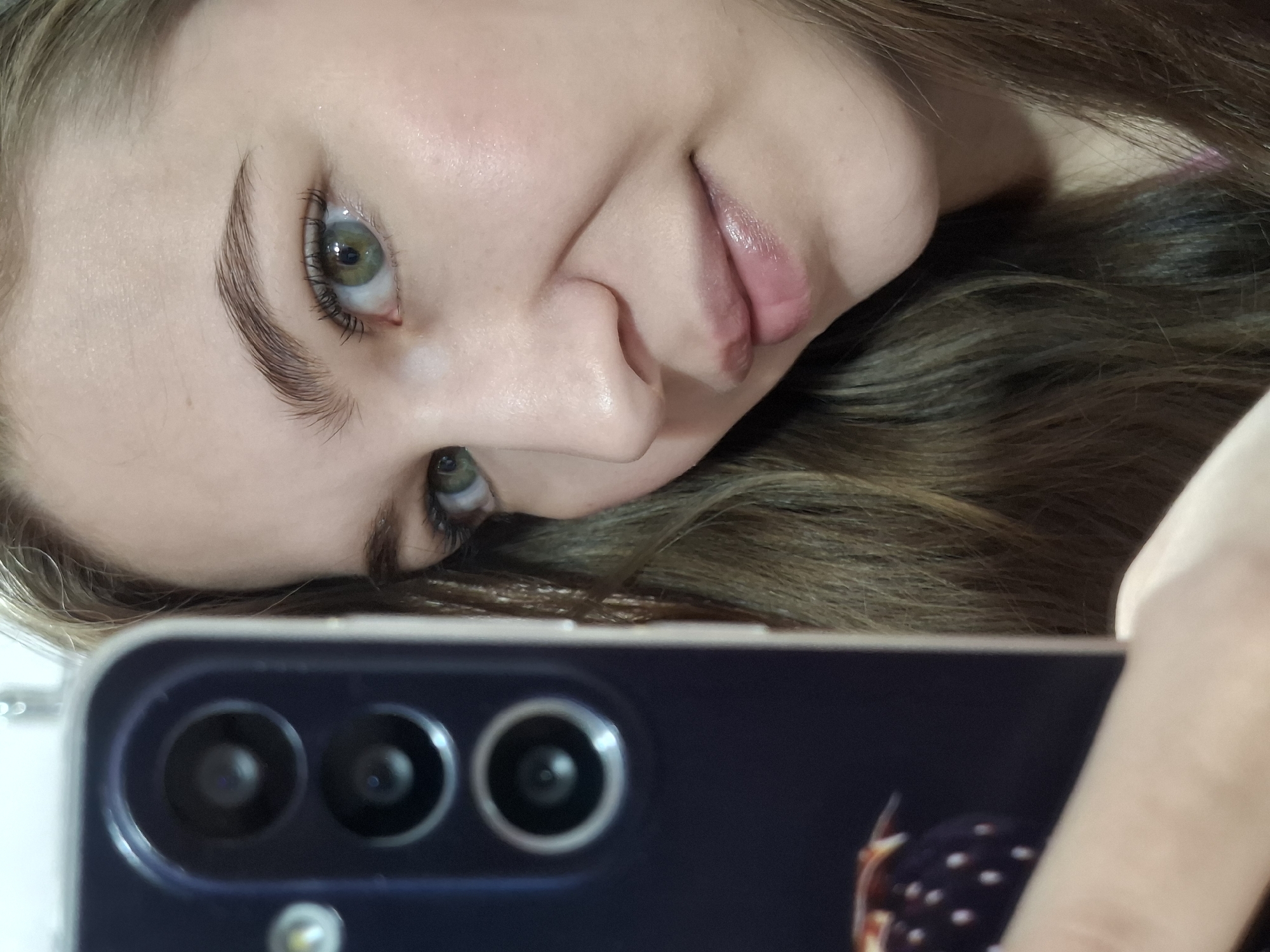 A young woman is holding a dark blue phone with three camera lenses to take a selfie. Her face is angled towards the camera. She has light skin, green eyes, and glossy lips. Her brown hair is slightly out of focus. The background is blurred, providing no clear identifiers. There is no evidence to indicate that the image was taken in Chaikovsky, Russia.