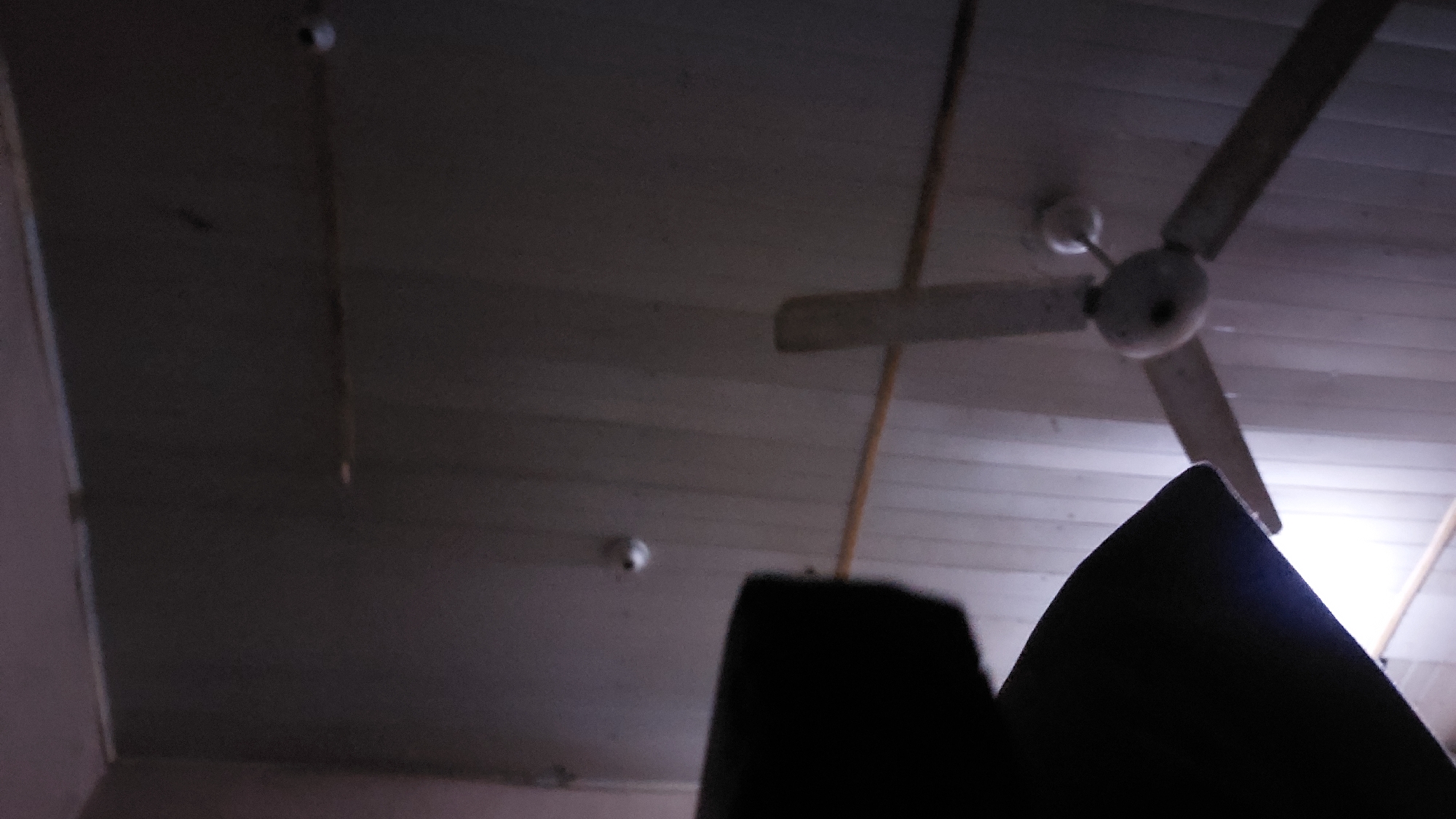 The image depicts a low-angle view of a ceiling with a wooden plank construction. A ceiling fan with three beige blades is visible, mounted centrally on the ceiling. Two small, white circular objects, possibly smoke detectors or lights, are also affixed to the ceiling. The scene appears to be indoors, with the lighting suggesting it might be at night or in a dimly lit environment. The lower part of the image is obscured by dark, indistinct shapes, likely furniture or other objects in the room, silhouetted against the ceiling. There are no people visible in the image. The location is identified as a property in New Nyanya, Nigeria.