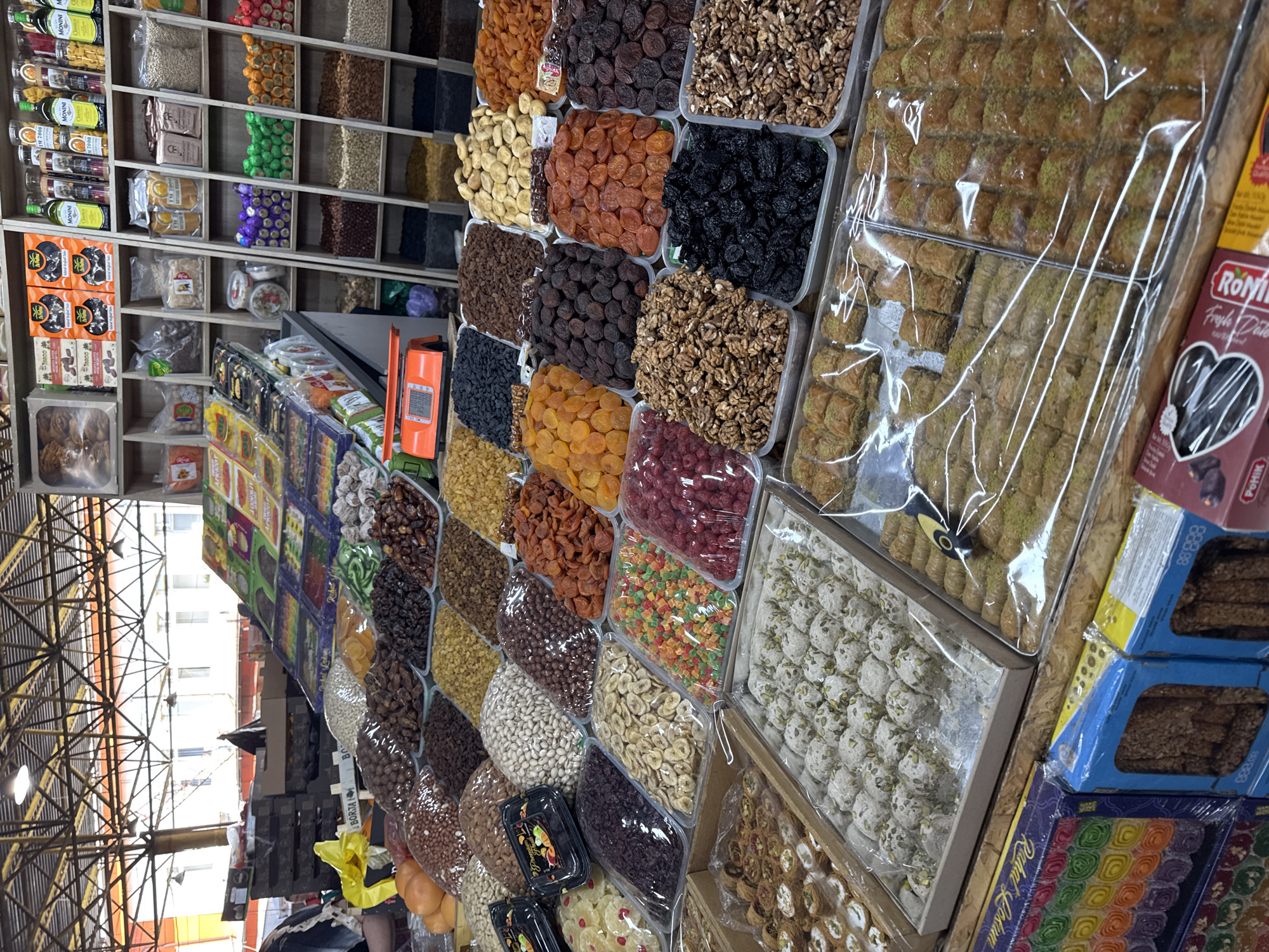 The image shows a market stall in Odesa, Ukraine, filled with a variety of dried fruits, nuts, and sweets. The stall is well-stocked with numerous plastic containers and boxes showcasing different types of products. In the foreground, there are trays of halva, baklava, and other confections. Further back, rows of nuts like walnuts and almonds are visible, alongside dried fruits such as apricots, dates, and prunes. Boxes of colorful Turkish delight are also displayed. The stall appears to be part of a larger market with a metal roof structure visible in the background. A person's arm is visible on the left side of the image, suggesting they are either a vendor or a customer. The overall impression is one of abundance and variety in a traditional market setting.