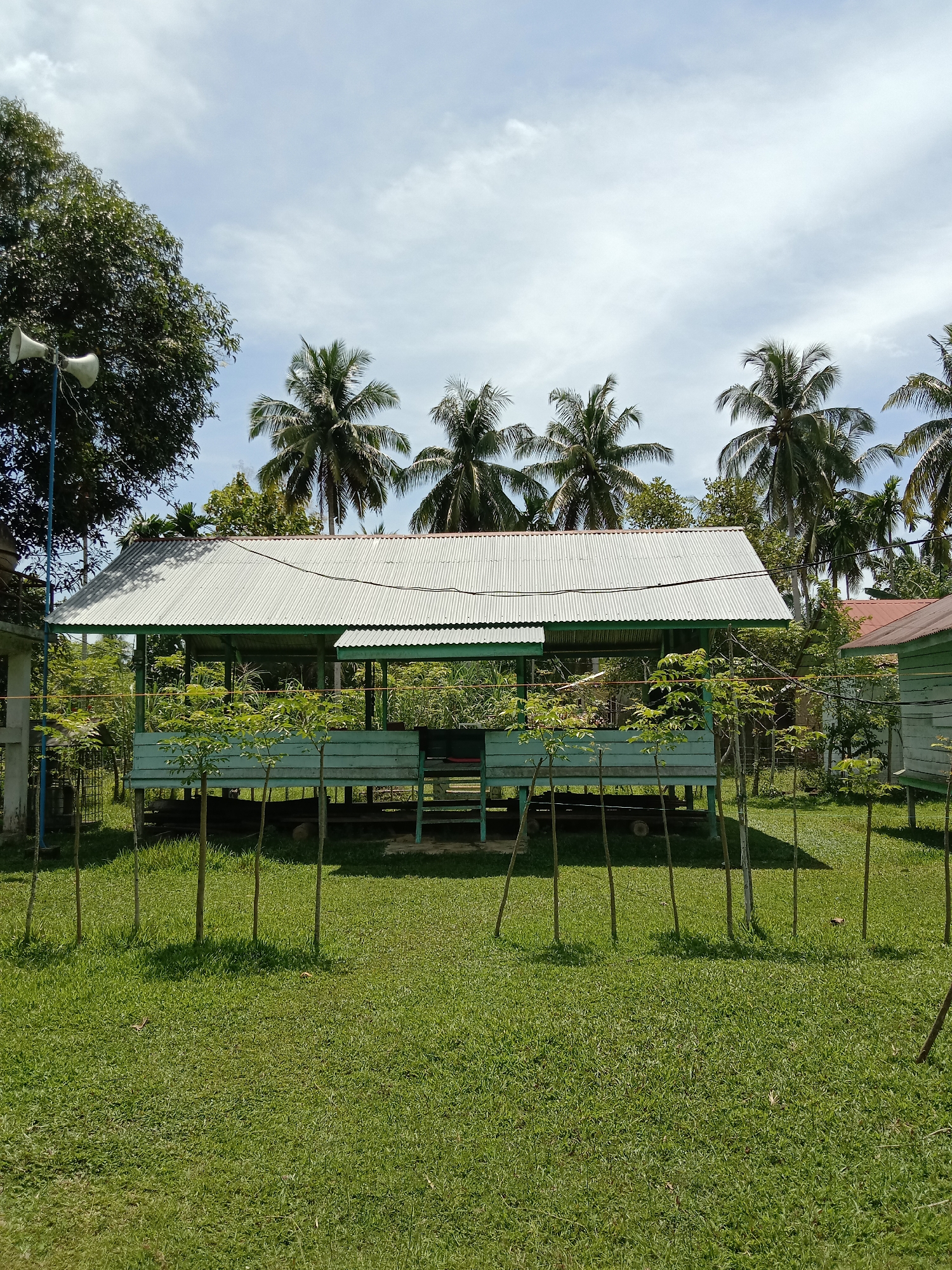The image shows a light blue, stilted structure with a corrugated metal roof, located in Blangkarieng, Indonesia. The structure is surrounded by a low fence of thin wooden stakes. The foreground is a green lawn. In the background, there are palm trees and other buildings. A loudspeaker is mounted on a pole to the left of the structure. The sky is partly cloudy.