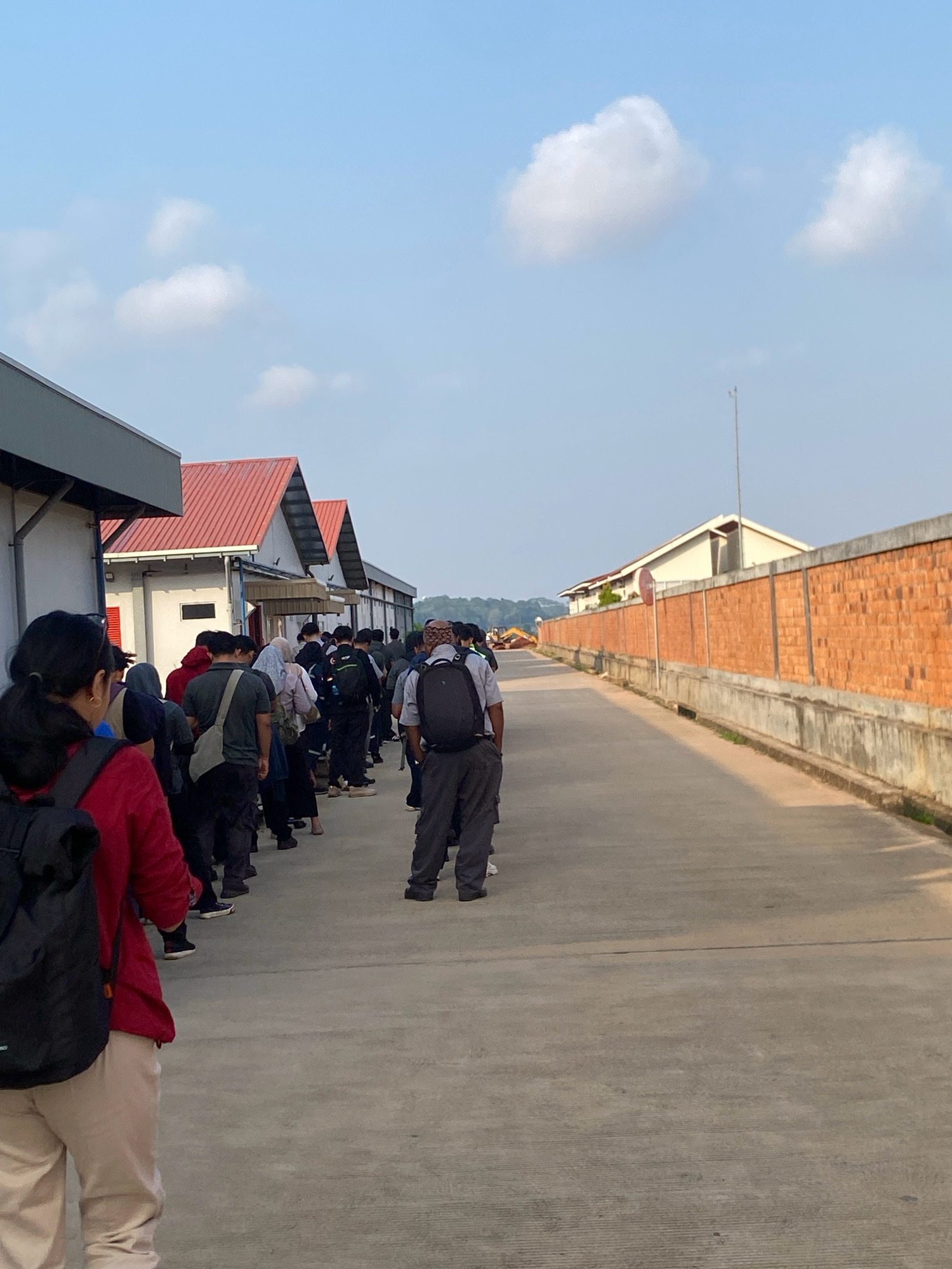 A group of people are lined up on a paved walkway in front of industrial buildings. The buildings have white walls and red-tiled roofs, with one building featuring a prominent grey overhang. To the right of the walkway is a tall brick wall. The sky is clear with scattered white clouds. The people are diverse in age and gender, many are wearing backpacks. Some individuals are wearing headscarves. The overall impression is an outdoor scene in a commercial or industrial area, possibly a factory or warehouse complex, located in Batam, Indonesia.
