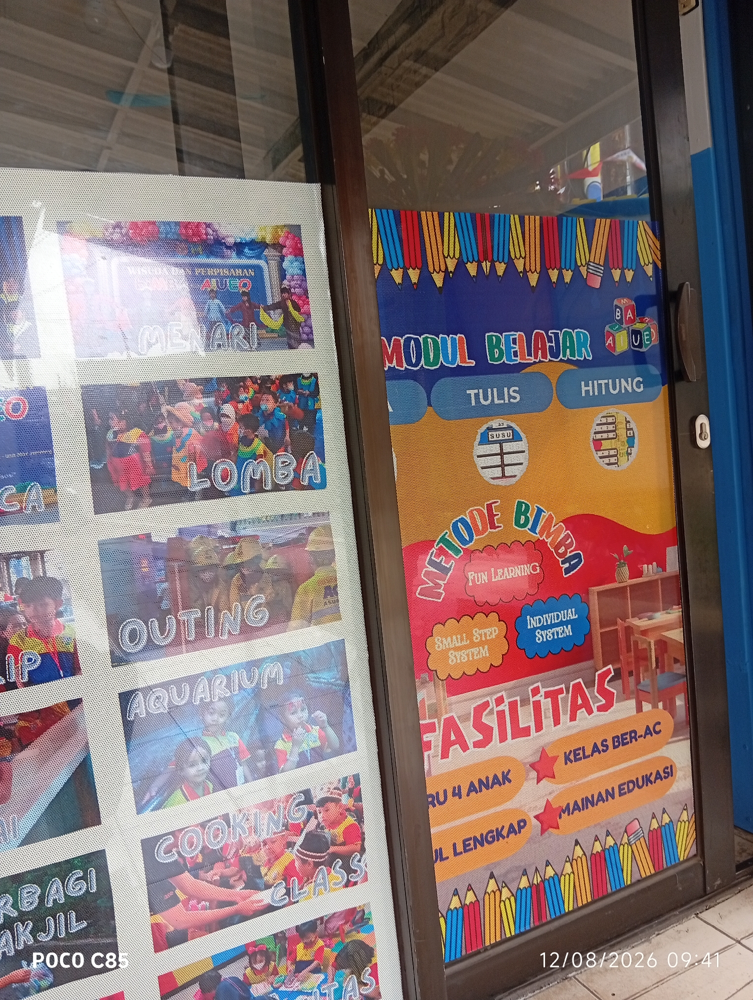 The image shows a storefront with two main sections displaying posters. The left side has a grid of smaller posters with Indonesian text and images. Visible titles include "MENARI" (Dancing), "LOMBA" (Competition), "OUTING", "AQUARIUM", "COOKING CLASS", and "BERBAGI AKJIL". The right side features a larger poster with the title "MODUL BELAJAR" (Learning Module) at the top, followed by "TULIS" (Write) and "HITUNG" (Count). Below this, there are sections for "METODE BIMBA" (BIMBA Method) with "FUN LEARNING", "SMALL STEP SYSTEM", and "INDIVIDUAL SYSTEM". Further down, the word "FASILITAS" (Facilities) is prominently displayed, with sub-sections like "KELAS BER-AC" (Air-conditioned Class), "RU 4 ANAK" (Room for 4 Children), and "MAINAN EDUKASI" (Educational Toys). The bottom right corner shows a date and time stamp: "12/08/2026 09:41". The background on the left appears to be a building interior with reflective surfaces, possibly a window. The right side is a glass door, and through it, a shelf unit with wooden chairs and tables is visible, suggesting an educational or children's facility. The overall context points to a kindergarten or early childhood education center. The location is identified as South Tangerang, Indonesia, based on the common use of Indonesian language in such establishments and the regional context.