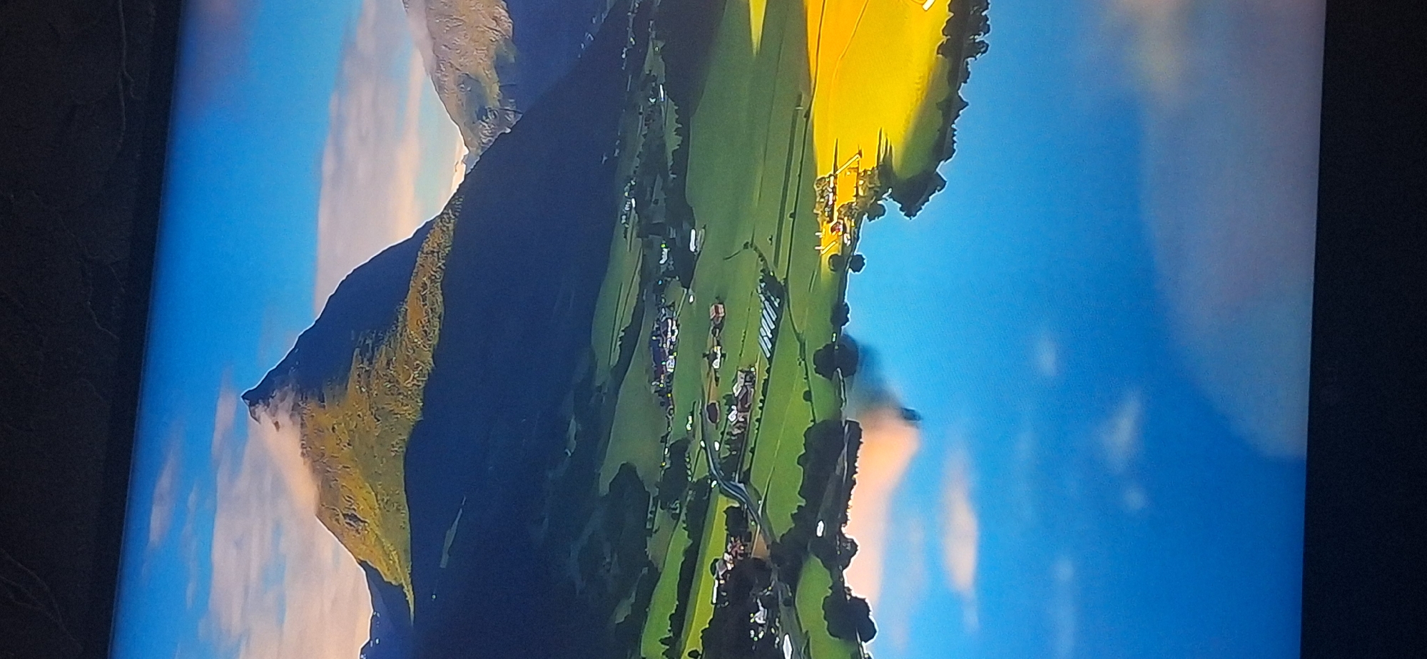 The image depicts a picturesque mountainous landscape with a serene lake in the foreground. A sharp, pyramid-shaped mountain dominates the background, its peak partially shrouded in wispy clouds. The mountain's slopes are covered in lush green vegetation, transitioning to darker shadows on the left side, indicating the direction of the light source. Below the mountain, rolling green hills are dotted with a small cluster of houses and what appear to be farm buildings, suggesting a rural settlement. A road winds its way through the landscape, connecting different parts of the village.
The foreground is occupied by a calm, reflective lake that mirrors the sky, the mountain, and the surrounding landscape. The water's surface is a deep blue, interspersed with reflections of white clouds and the warm hues of the sky. The right side of the lake is bordered by a field of vibrant yellow, possibly a field of crops or flowers, and a small group of buildings.
The scene is bathed in the soft, warm light of either sunrise or sunset, casting long shadows and creating a peaceful, idyllic atmosphere. The overall impression is one of natural beauty and tranquility.
There is no discernible connection to the city of Bila Tserkva, Ukraine, as the image portrays a mountainous, possibly European, rural or alpine environment, which is not characteristic of Bila Tserkva. The provided image does not contain any elements that would place it in that specific location.