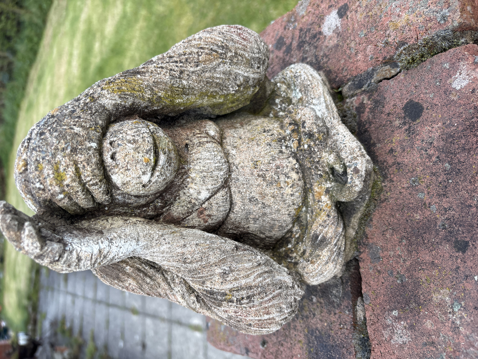 The image shows a weathered stone sculpture of a sleeping child, possibly an angel or cherub, resting on a weathered brick surface. The sculpture is light grey with visible surface texture and minor moss growth. The child is depicted lying on its side, with its head supported by its arm. The brick surface is red-brown with visible moss and discoloration, suggesting age and exposure to the elements. Background includes grass, bricks, and pavement suggesting that this image was taken in a garden. The sculpture is in Shrewsbury, United Kingdom.