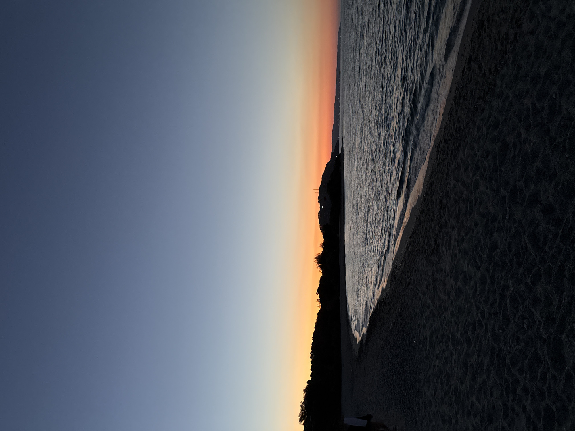 The image depicts a twilight scene at a beach in the Kassandra Municipal Unit, Greece. The sky transitions from a deep blue at the top to a vibrant orange and pink horizon just above the silhouetted coastline. Dark, undulating sand forms the foreground, with gentle waves lapping at the shore. The sea is a deep, dark blue, reflecting the colors of the sky. Silhouettes of trees and structures are visible on the distant hills, which are also cloaked in shadow. A single, indistinct figure is visible on the left side of the frame, walking on the beach. The overall impression is one of tranquility and the end of a day.