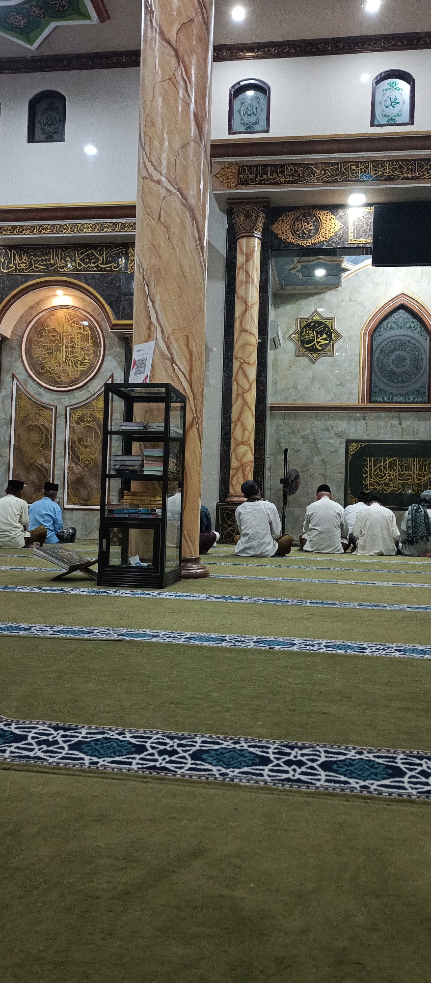 The image depicts the interior of a mosque in Arjasa, Indonesia, likely during a prayer session. Several men are seated on a green carpet with decorative borders, facing away from the camera, in a prayer posture. The mosque features ornate Islamic calligraphy on its walls and decorative columns. A tall, marble-patterned pillar stands prominently in the foreground. A black shelf unit containing books is positioned to the left, and a stand holding a religious book is visible near the pillar. The environment appears to be indoors, with artificial lighting.