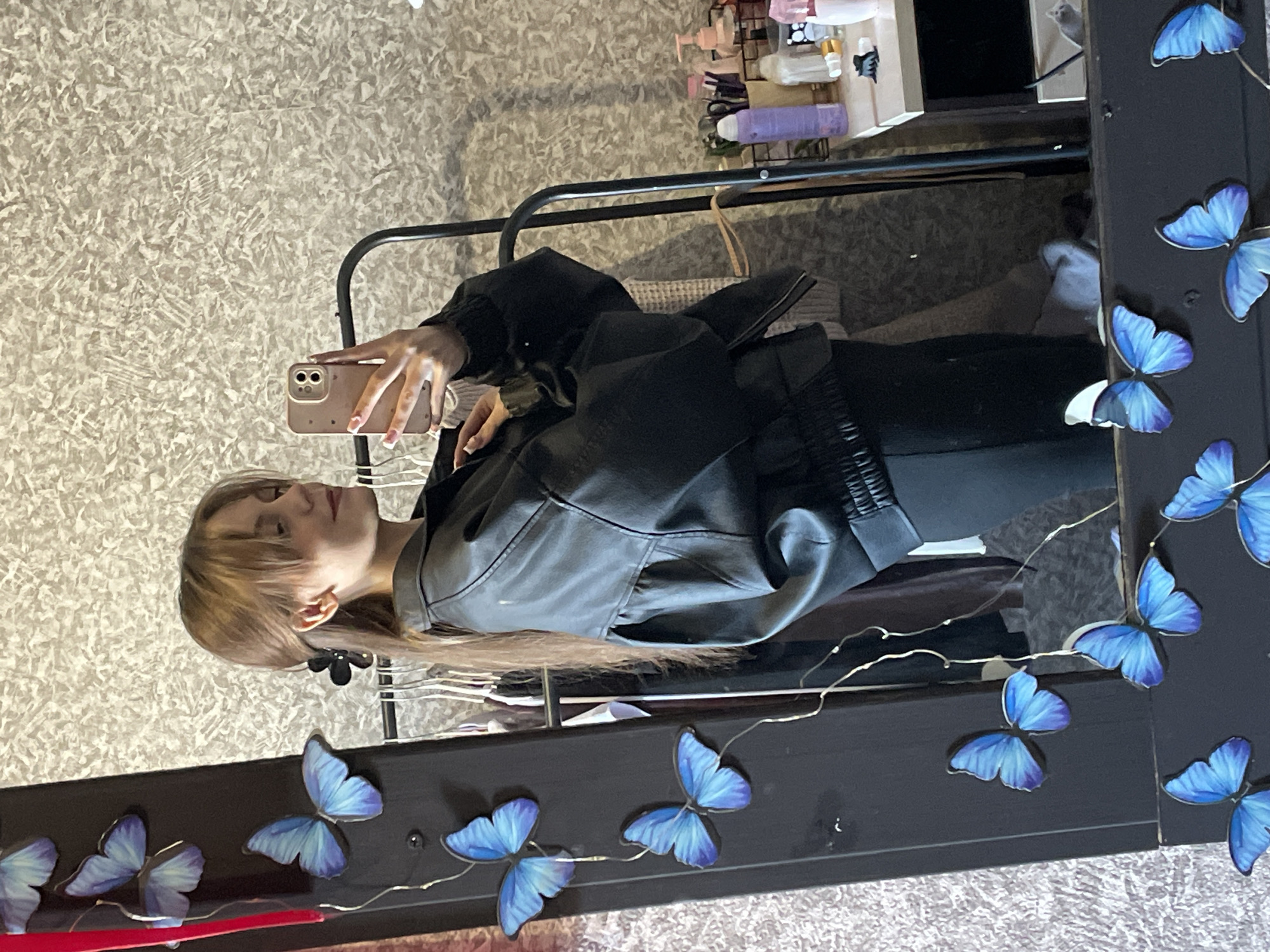 An indoor mirror selfie taken in the city of Фоки, Russia, depicts a female subject from the chest up. The female has light brown hair styled with bangs and a portion pulled back with a black claw clip. She is wearing a black bomber-style jacket over a dark garment and black pants. Her right hand, with light-colored fingernails featuring white tips, holds a light-colored smartphone with a triple camera array, aimed at the mirror. She is slightly angled to her right with her head tilted, and a subtle smile is visible.
The subject is reflected in a tall, dark-framed mirror. The mirror's frame is adorned with multiple blue decorative butterflies of varying sizes and a string of delicate fairy lights. A vertical red element is present on the left edge of the mirror frame. Reflected in the background behind the subject is a light-colored, textured wallpaper with an abstract, mottled pattern. To the viewer's right in the reflection, a black metal clothing rack with garments is visible. To the viewer's left in the reflection, a light-colored shelf or table holds an assortment of personal care items, including bottles, pump dispensers, and brushes.