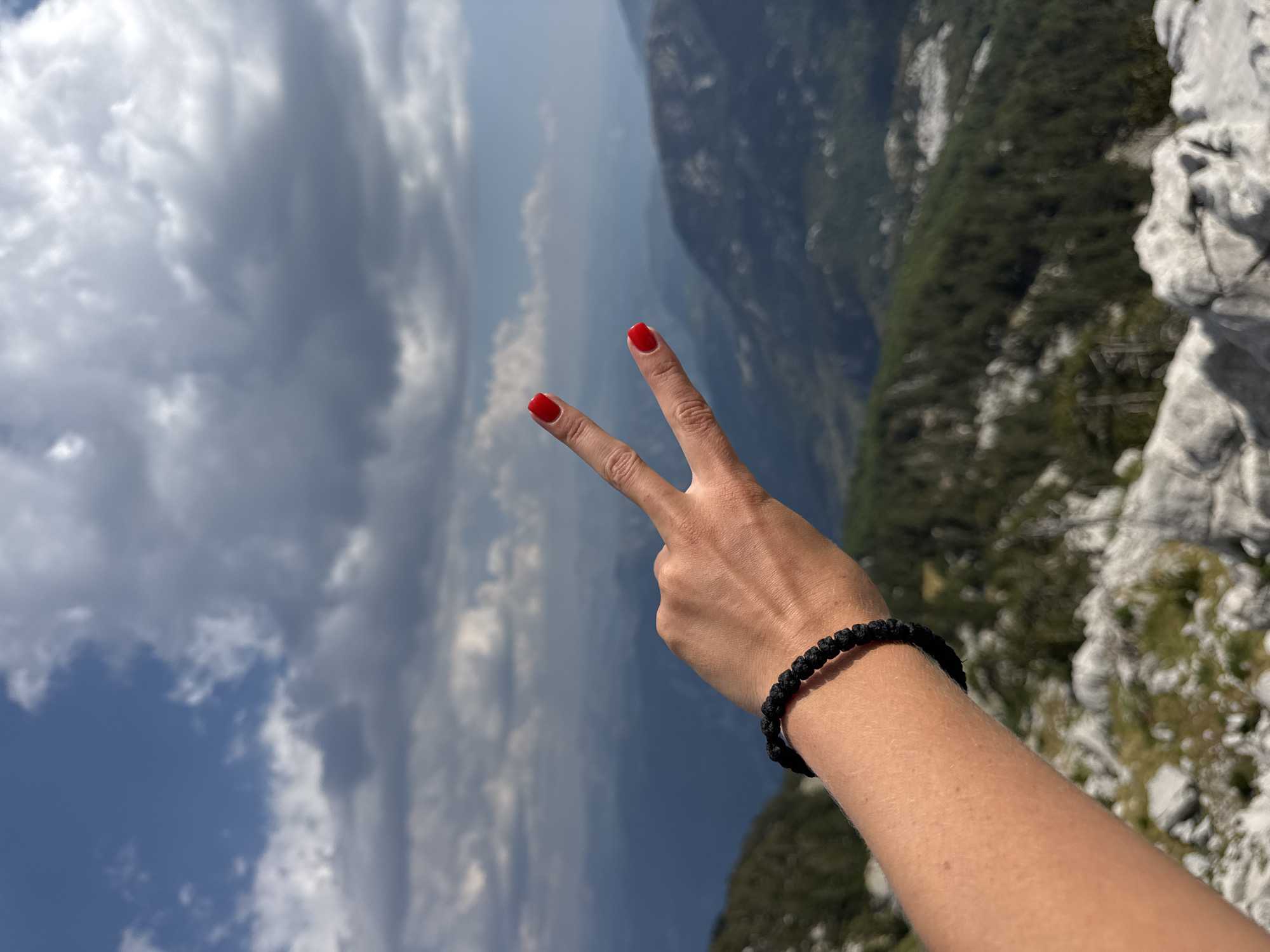 A person's hand, with red nail polish, forms a peace sign against a backdrop of a mountainous landscape. The hand is adorned with a black beaded bracelet. The environment appears to be Mrtvo Duboko, Montenegro, characterized by rugged terrain with rocky outcrops and sparse vegetation. The sky above is partially cloudy, with patches of blue visible.
