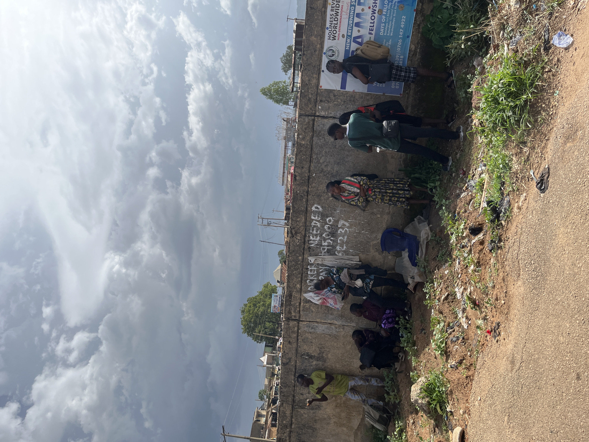 In Jos, Nigeria, a group of people are gathered near a concrete wall on a roadside. The sky is filled with dramatic, grey clouds. Several individuals are standing and sitting along the base of the wall, some appear to be waiting. One person in a lime green shirt stands with their hands up, looking to the left. Others are seated, with a few carrying bags. Graffiti or writing is visible on the wall, including the words "WORKERS NEEDED" and numbers. A large banner with text, possibly religious or organizational, is affixed to the wall on the right side of the frame. The ground in front of the wall is littered with debris, including plastic bags, scattered trash, and some vegetation. The road surface is visible in the foreground. Beyond the wall, buildings and trees are visible, indicating an urban or semi-urban environment.