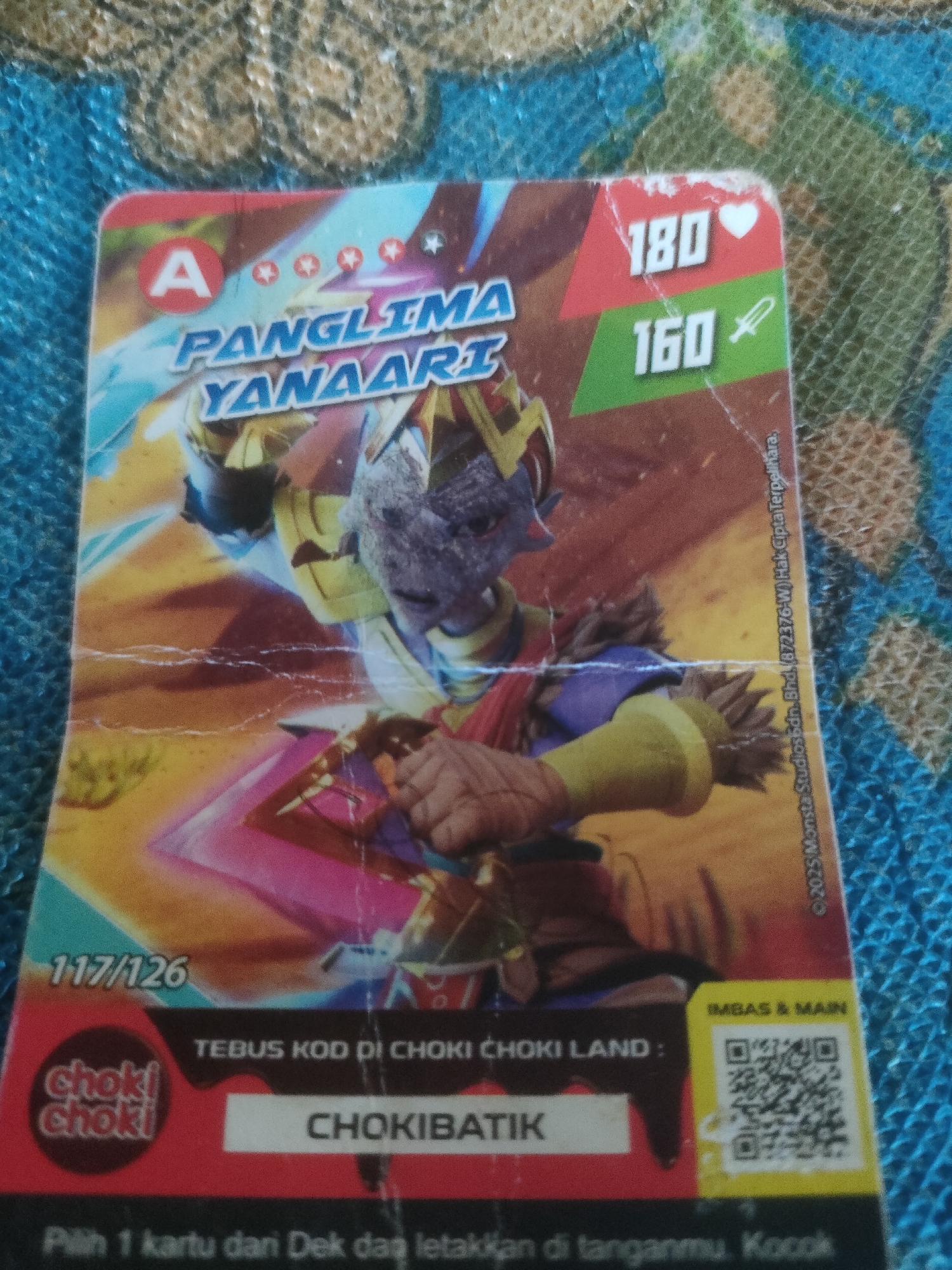 The image is a close-up, overhead shot of a trading card featuring a character from a cartoon or video game, likely "Bimbi" from "Choki Choki Land" based on the text on the card. The card has a character named "Panglima Yanaart" with a power level of 180 and a sword symbol with a level of 160. The card is worn and creased, suggesting it has been handled frequently. The background is a blue, woven fabric with a green and tan pattern, possibly a tablecloth or a mat. Text on the card includes "CHOKIBATIK" and instructions to "TEBUS KOD DI CHOKI LAND". The copyright information at the bottom right reads "© 2023 Monsta Studios Sdn. Bhd. (872376-W) Hak Cipta Terpelihara". The card also displays a QR code under the heading "IMBAS & MAIN". There are no indications in the image to suggest a specific location within the city of Padang Dalom, Indonesia.