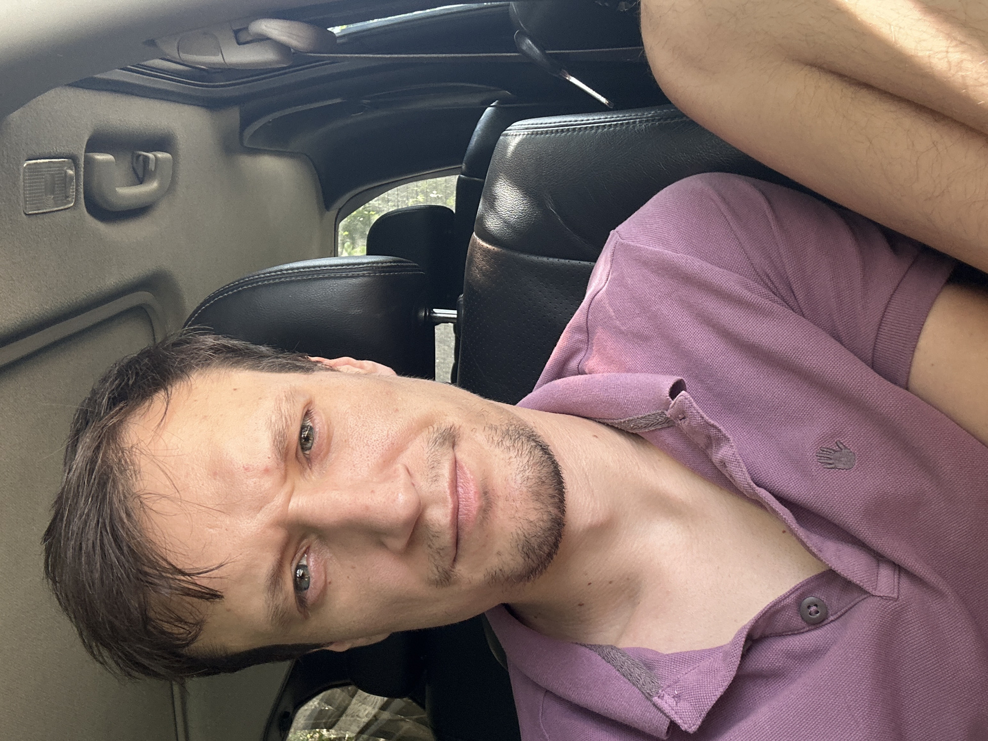 A man with short brown hair and light stubble is seated in the driver's seat of a car. He is wearing a purple polo shirt. The interior of the car is visible, including black leather seats and the ceiling. Sunlight streams in from the side, casting shadows on the interior. The man's expression is serious, with his brow furrowed. The background outside the car window shows foliage, suggesting a location with trees. It is not possible to determine the specific location within the city of Makiv, Ukraine, from this image.