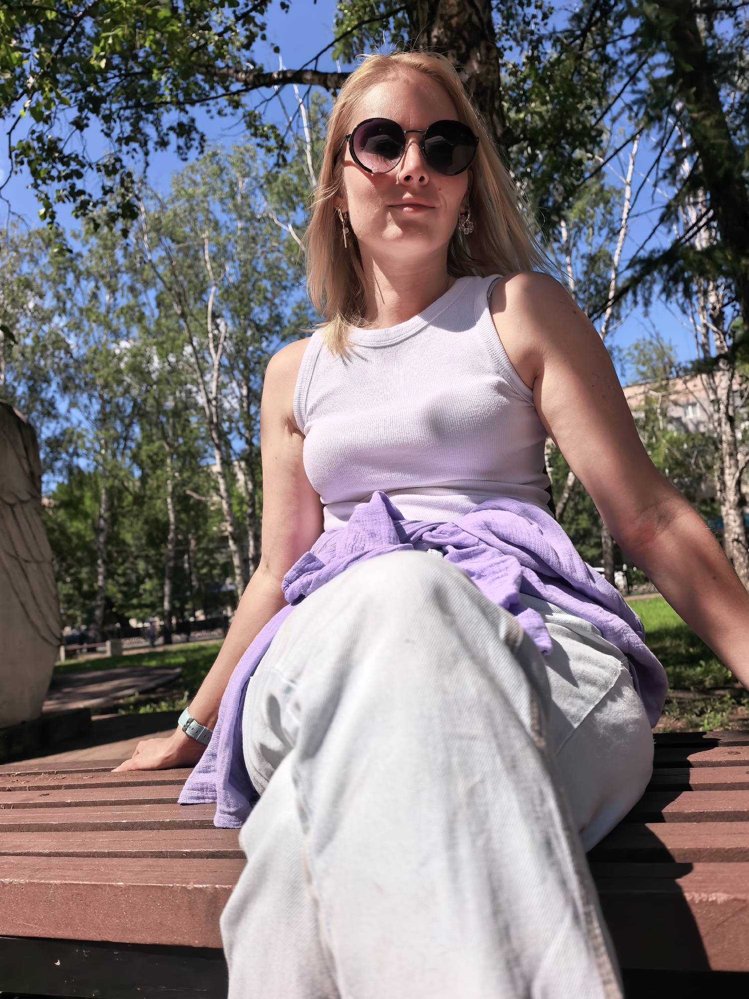 A woman with blonde hair and sunglasses sits on a wooden bench in a park, with trees and buildings in the background. She is wearing a white tank top, light-colored pants, and a purple sweater tied around her waist. A light blue watch is visible on her left wrist. The woman is positioned in the foreground, with her legs extending towards the camera. The scene appears to be in Sterlitamak, Russia, based on the provided context, and it is daytime with clear skies.
