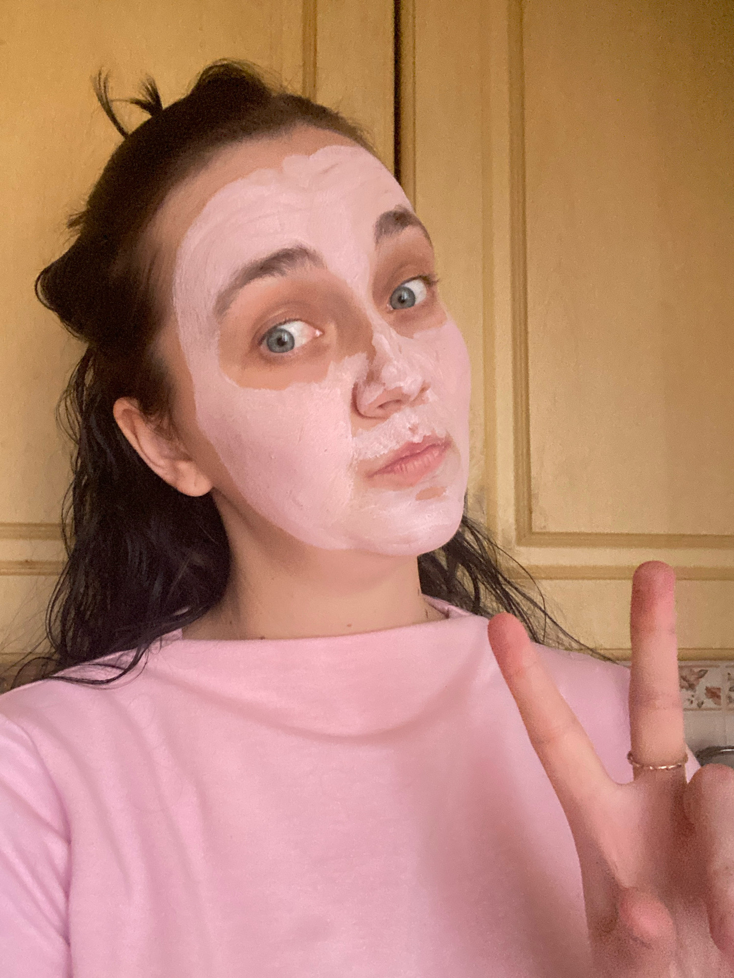 A young woman with dark, wet hair pulled up into a messy bun is pictured from the chest up, wearing a pink t-shirt. She has a pink face mask on, covering most of her face except for her eyes, eyebrows, and lips. Her eyes are wide open and looking at the camera. She is holding up two fingers in a peace sign with her right hand, wearing a small ring on her ring finger. The background appears to be a kitchen with light brown wooden cabinets and a tiled backsplash. The lighting is natural, coming from the right. The woman is likely in her home.