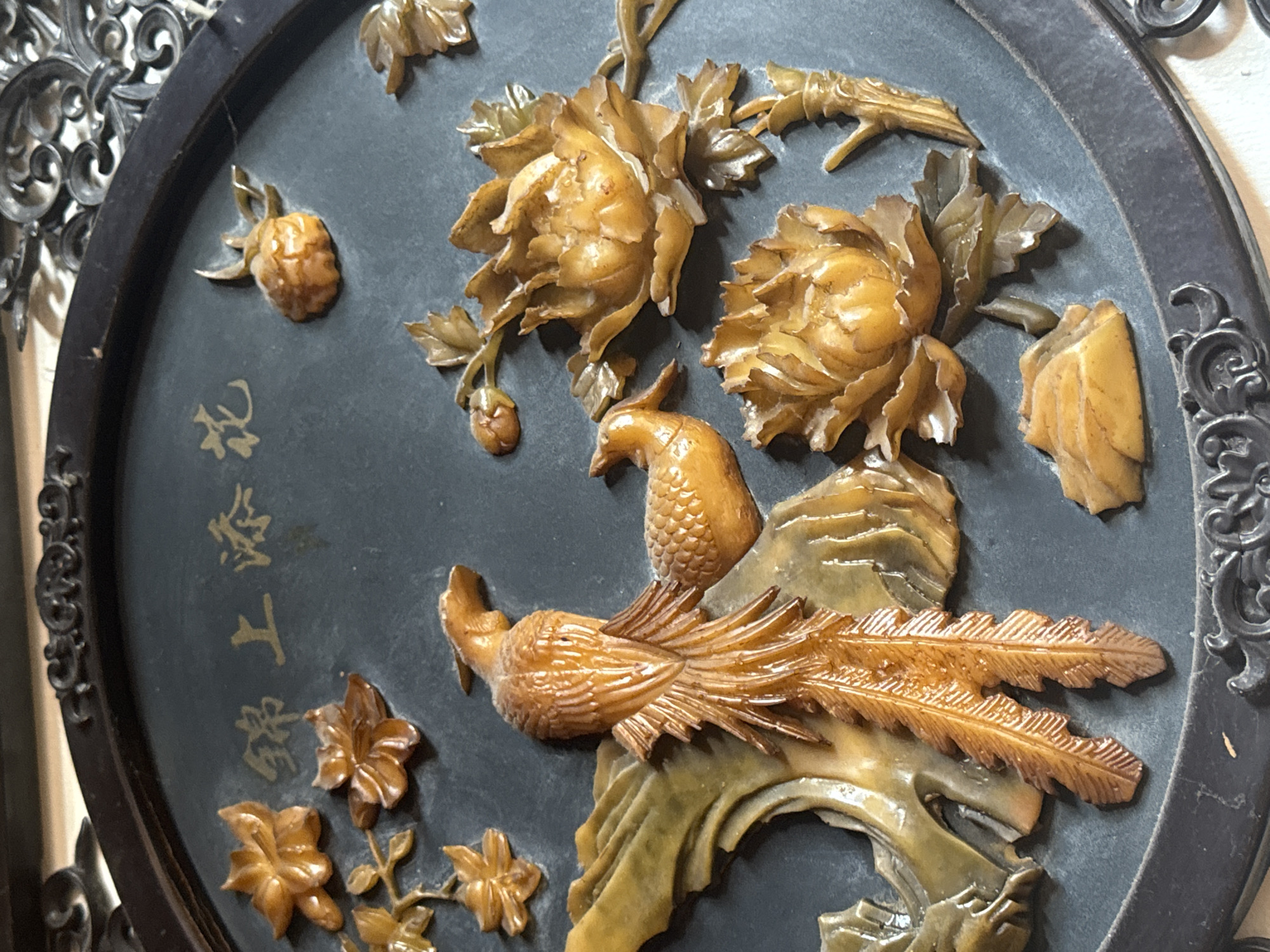A decorative circular plaque, likely made of carved stone, resin, or similar material, is depicted in a close-up shot. The plaque features intricate relief carvings of two stylized birds, resembling peacocks or phoenixes, among various flowers and foliage, all rendered in a yellowish-brown, translucent material that suggests jade or soapstone. The background of the plaque is a dark, matte gray or black.
The larger bird is positioned towards the bottom right, with a long, flowing tail detailed with finely carved feathers. Its head is turned towards the left. A smaller bird is situated above the larger one, with its head also facing left. Surrounding the birds are several large, full-blossomed flowers, possibly peonies, characterized by layered petals. Smaller leaves, buds, and a berry-like cluster are also integrated into the design. The carvings exhibit a traditional East Asian aesthetic.
On the left side of the dark background, golden Chinese characters are inscribed vertically. The visible text reads "献新工艺" (xiàn xīn gōng yì), which translates to "Offering New Craftsmanship" or "Presenting New Artistry". The plaque is framed by an ornate, dark-colored rim with intricate scrollwork, suggesting it is either a standalone decorative piece or part of a larger furnishing. The lighting highlights the raised carvings, creating shadows that emphasize their three-dimensional quality.