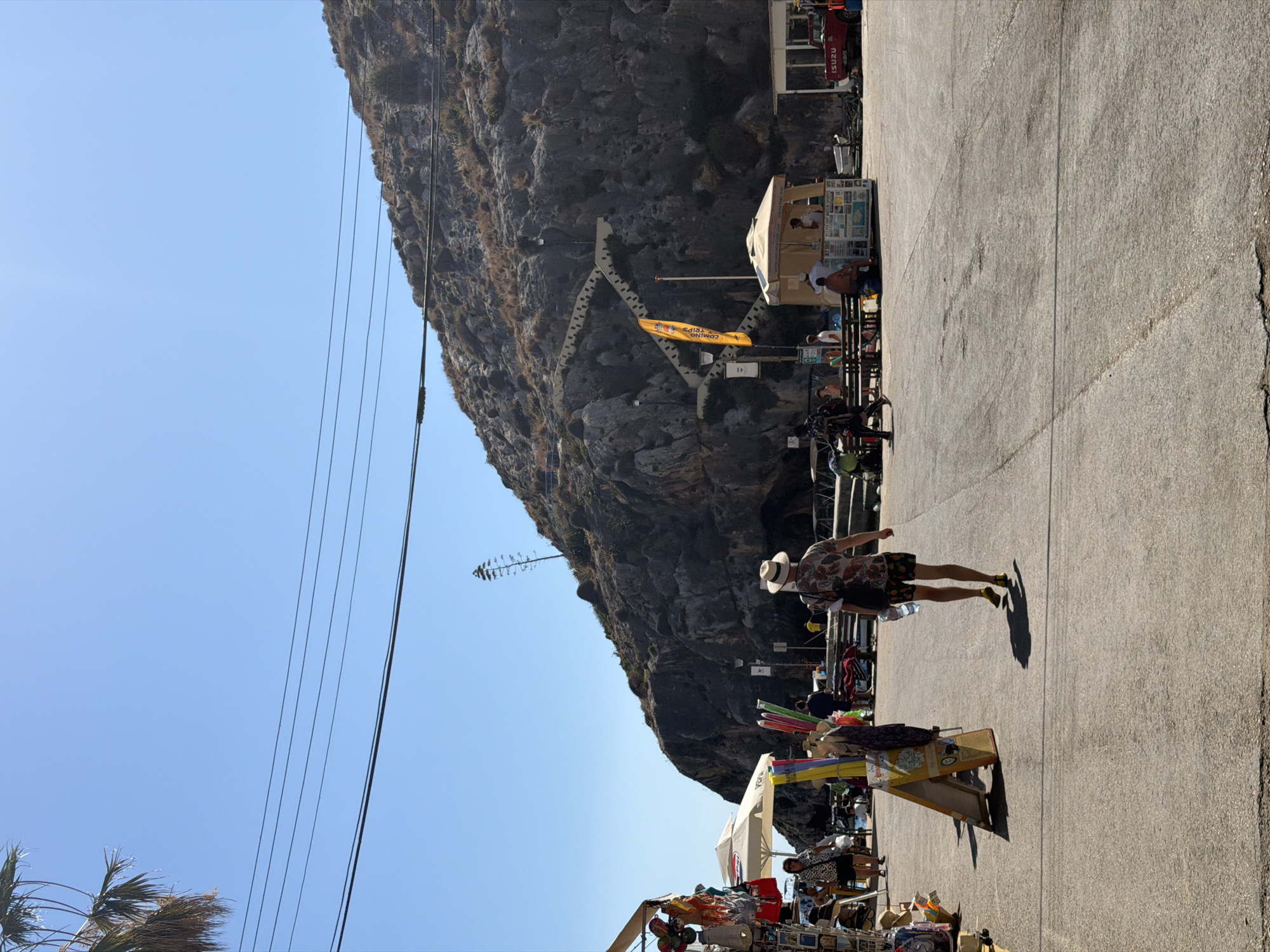 The image shows a street scene in Xlendi, Malta, with a large rocky hillside dominating the background. A winding staircase is visible on the hillside. In the foreground, a man wearing a hat and patterned shirt walks away from the camera. To the left, there are market stalls selling various items, including postcards and souvenirs, under a white umbrella. A kiosk is also visible, with people standing around it. A red Isuzu truck is parked on the right side of the image. The sky is clear and blue. The ground is paved with asphalt.