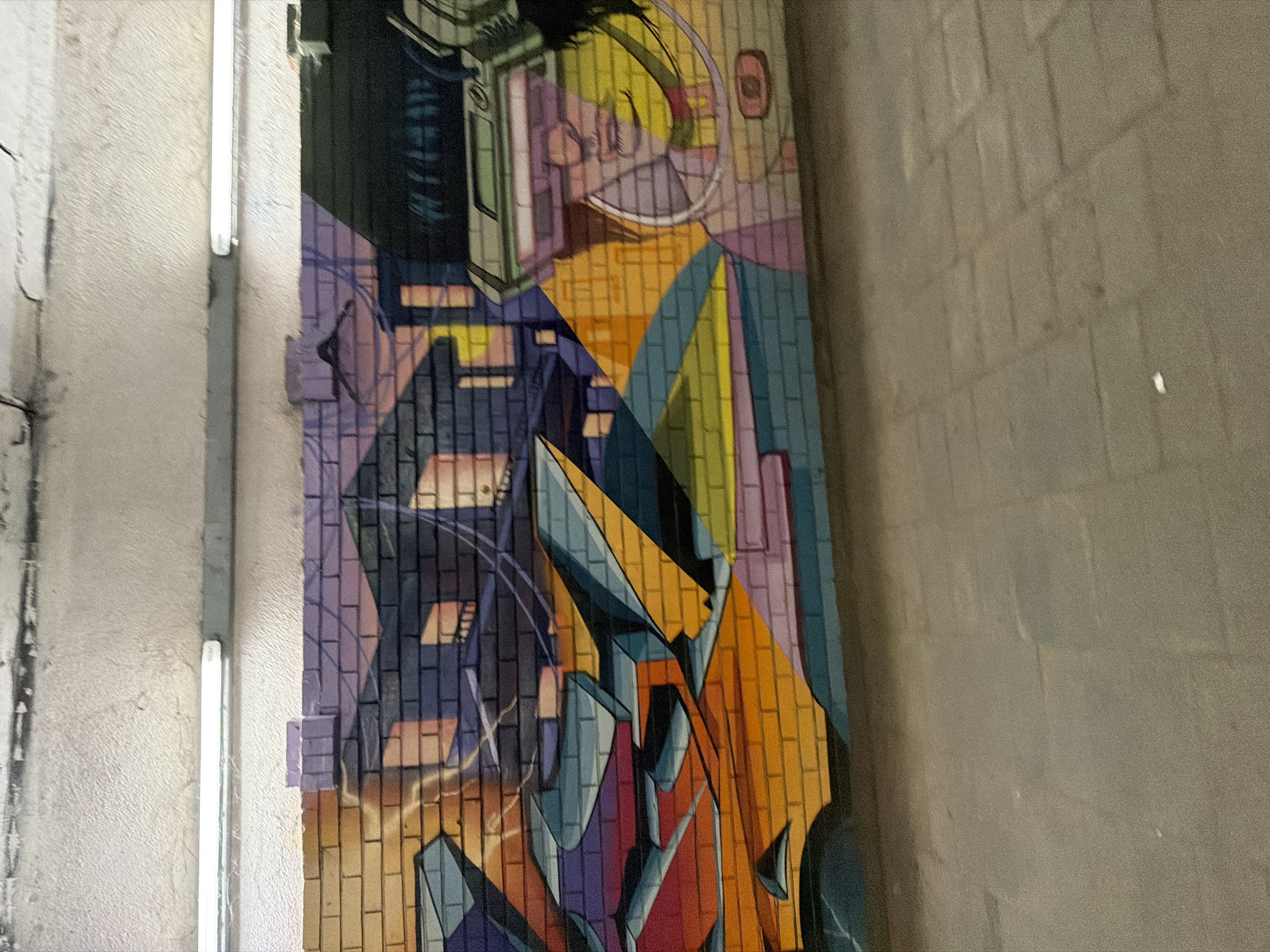 The image shows a graffiti mural on a brick wall in a city. The mural features abstract geometric shapes in bright colors, as well as a stylized depiction of a person wearing futuristic goggles. The background of the mural appears to be a cityscape at sunset, with silhouetted buildings and a gradient of purple and orange hues. The graffiti is illuminated by fluorescent lights mounted on the ceiling of what appears to be an underpass or tunnel. The ground in front of the wall is paved with concrete blocks. The overall impression is one of urban art in a public space.