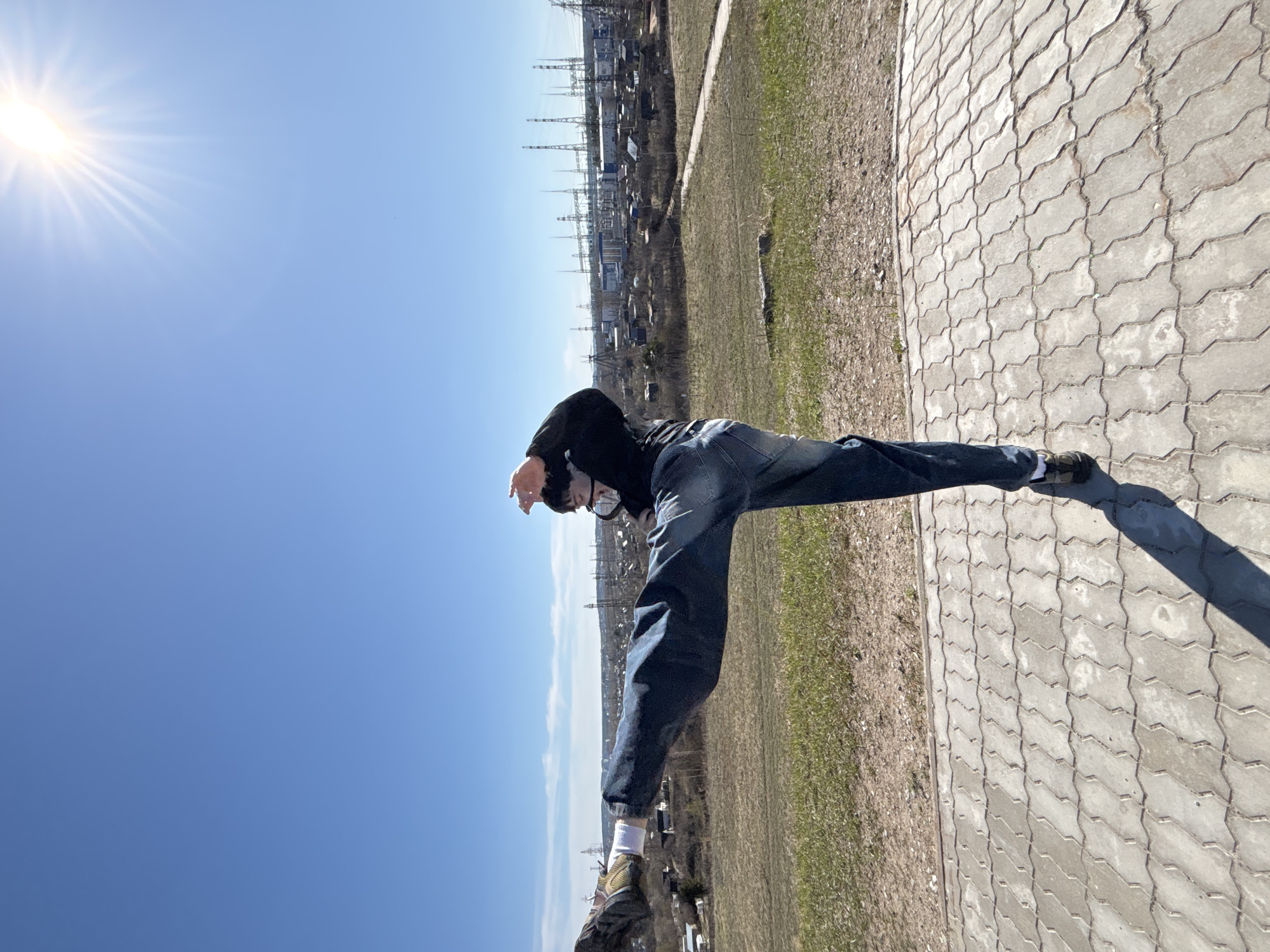 A person wearing jeans, a black hoodie, and a face mask is standing on a paved surface with a grassy area nearby. In the background, there are buildings and power lines, suggesting an urban area. The bright sun is visible in the upper left corner of the image. The setting appears to be in Chaikovsky, Russia, based on the presence of specific infrastructural details, but this cannot be confirmed without further information. The person is balancing or posing in a slightly unusual stance, possibly on the edge of a raised area.