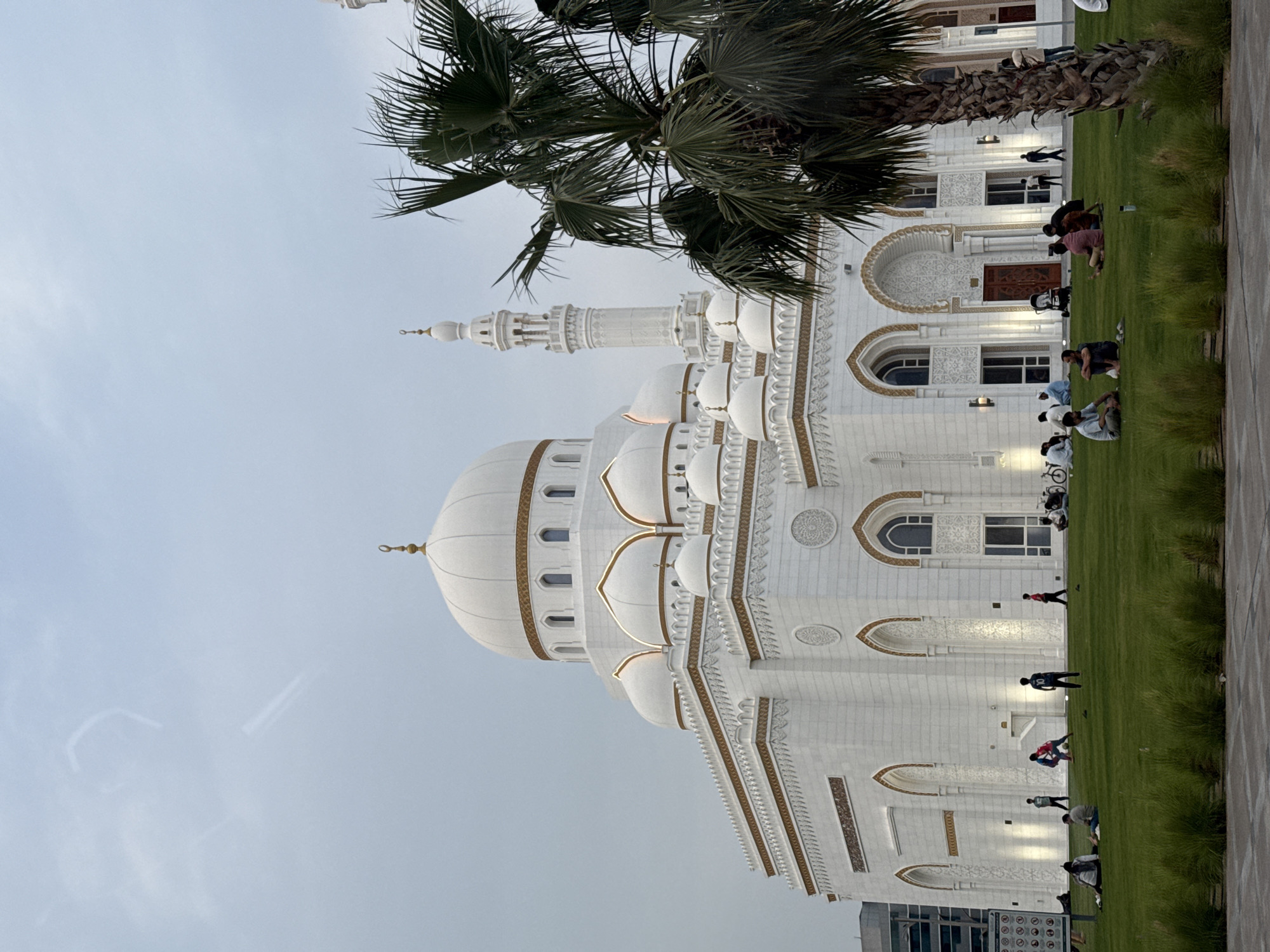 The image presents the Jumeirah Mosque in Dubai, United Arab Emirates, from a low vantage point. The mosque, characterized by its white facade and Islamic architectural elements, dominates the view. A palm tree is positioned in front of the mosque, partially obscuring the structure. Several people can be seen on the lawn adjacent to the mosque, some of whom appear to be praying or engaging in meditative practices. The sky is overcast, suggesting it might be early morning or late afternoon. A modern building can be seen in the background.