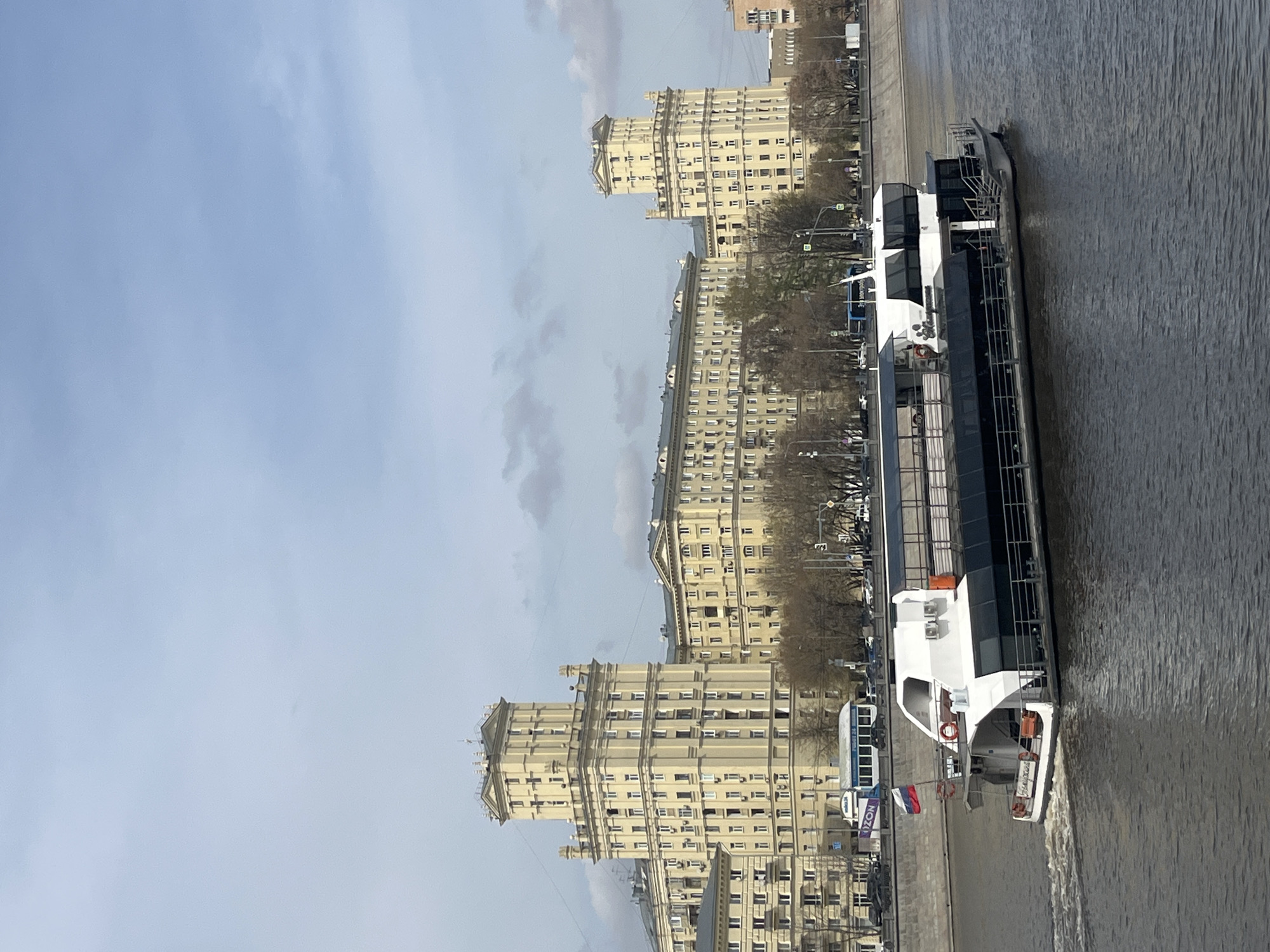 The image captures a daytime view of the Kotelnicheskaya Embankment Building in Moscow, Russia, situated alongside the Moskva River. A modern, multi-level riverboat is docked or moving along the river's edge, displaying the Russian flag. The architectural style of the building is Stalinist, evident in its grand design and symmetrical facade. The sky is overcast with scattered clouds. The environment appears urban, with trees lining the embankment.