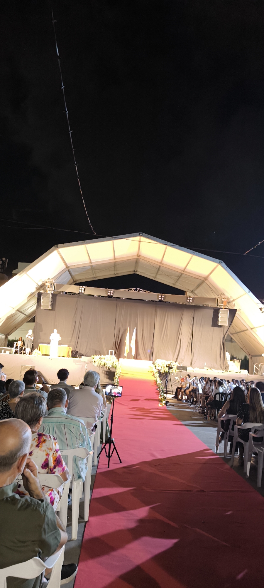 The image depicts an outdoor nighttime event at the town of Torreblanca, Spain, under a large, illuminated tent structure. A red carpet leads towards a stage draped with dark fabric. On the stage, two figures are visible, one appearing to be giving a speech or presentation, standing behind a table adorned with floral arrangements. To the right of the stage, a group of people are seated, possibly a musical ensemble. A significant number of spectators are seated in white chairs on either side of the red carpet, facing the stage. Many attendees are captured from the back, their attention directed towards the event. One spectator in the foreground is holding a tablet device, possibly recording or viewing the proceedings. The lighting from the tent casts a warm glow on the scene, and shadows stretch across the red carpet. The sky above is dark, with some faint wires or strings visible, likely part of the lighting or stage setup.