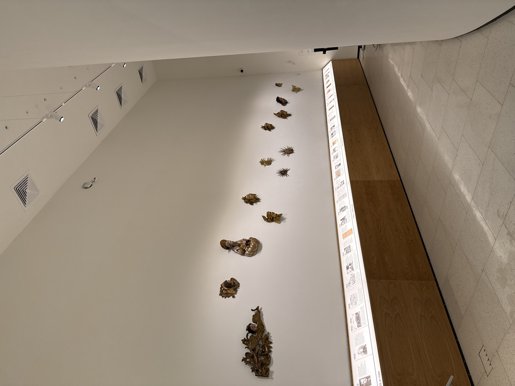 The image depicts an interior space, likely a museum or art gallery. It showcases a white wall with a series of gold-colored, decorative art pieces mounted vertically, ascending toward the top of the frame. Next to the art installation is a vertical panel with multiple small displays of text and images. The space is well-lit, and part of the floor is visible, with a light-colored stone pattern. The floor transitions to a wooden surface next to the display. The location is Perm, Russia.