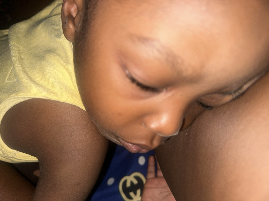 A close-up captures a baby sleeping, nestled against a darker-skinned arm or leg. The child's face dominates the frame with closed eyes and a peaceful expression. A yellow top peeks out from the left side, and a blue fabric with a blurred white and gold design appears below. The overall image conveys a sense of comfort and security.
