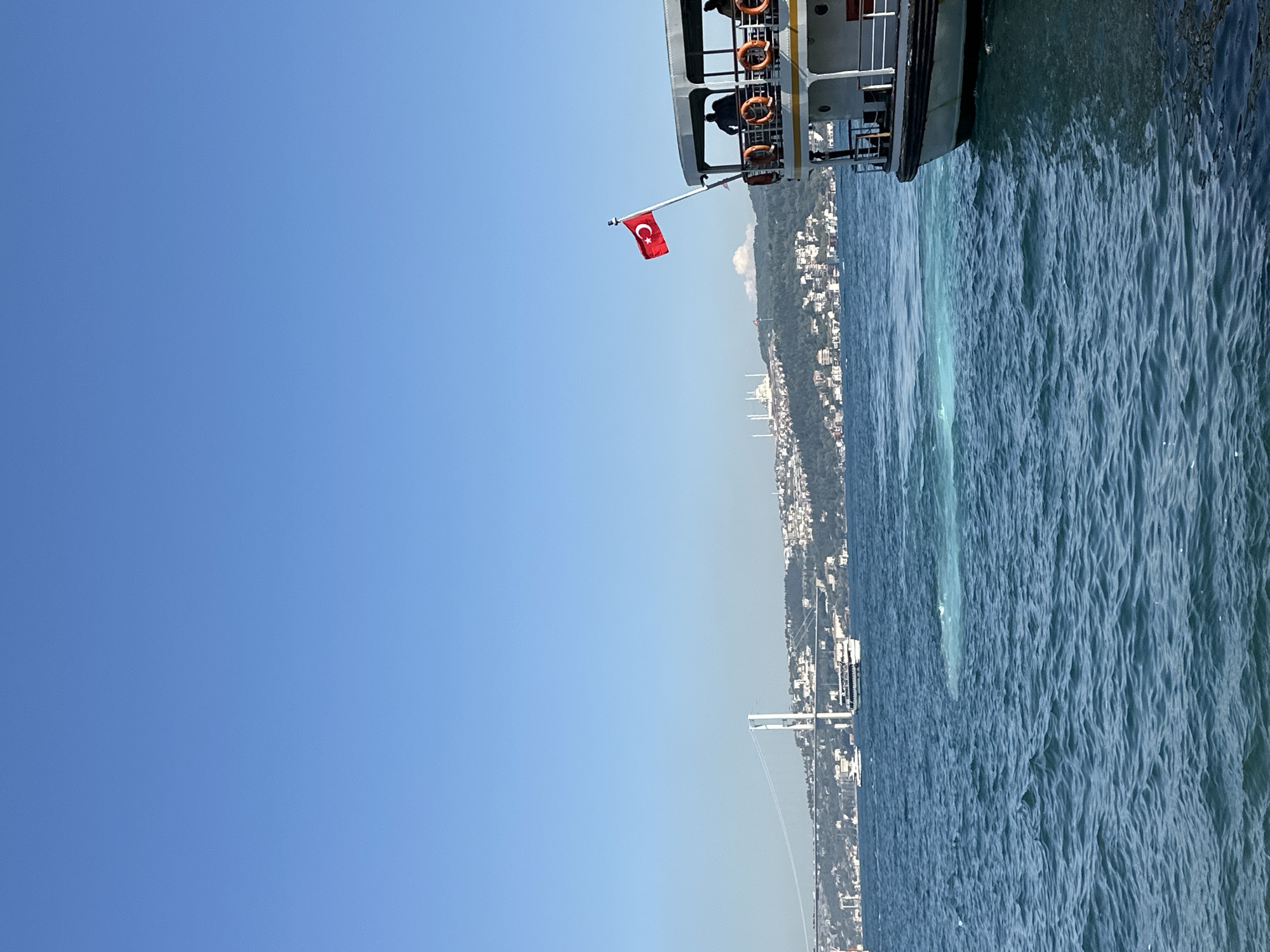 In Beşiktaş, Turkey, a ferry with a Turkish flag flies on its mast navigates the deep blue waters of the Bosphorus Strait. The ferry, a prominent subject in the foreground, displays life preservers on its railing and a few passengers can be seen on its deck. In the middle ground, another boat is visible on the water. The background features a cityscape nestled on a green, hilly terrain, with a notable white suspension bridge spanning across the strait. The sky is clear and blue.