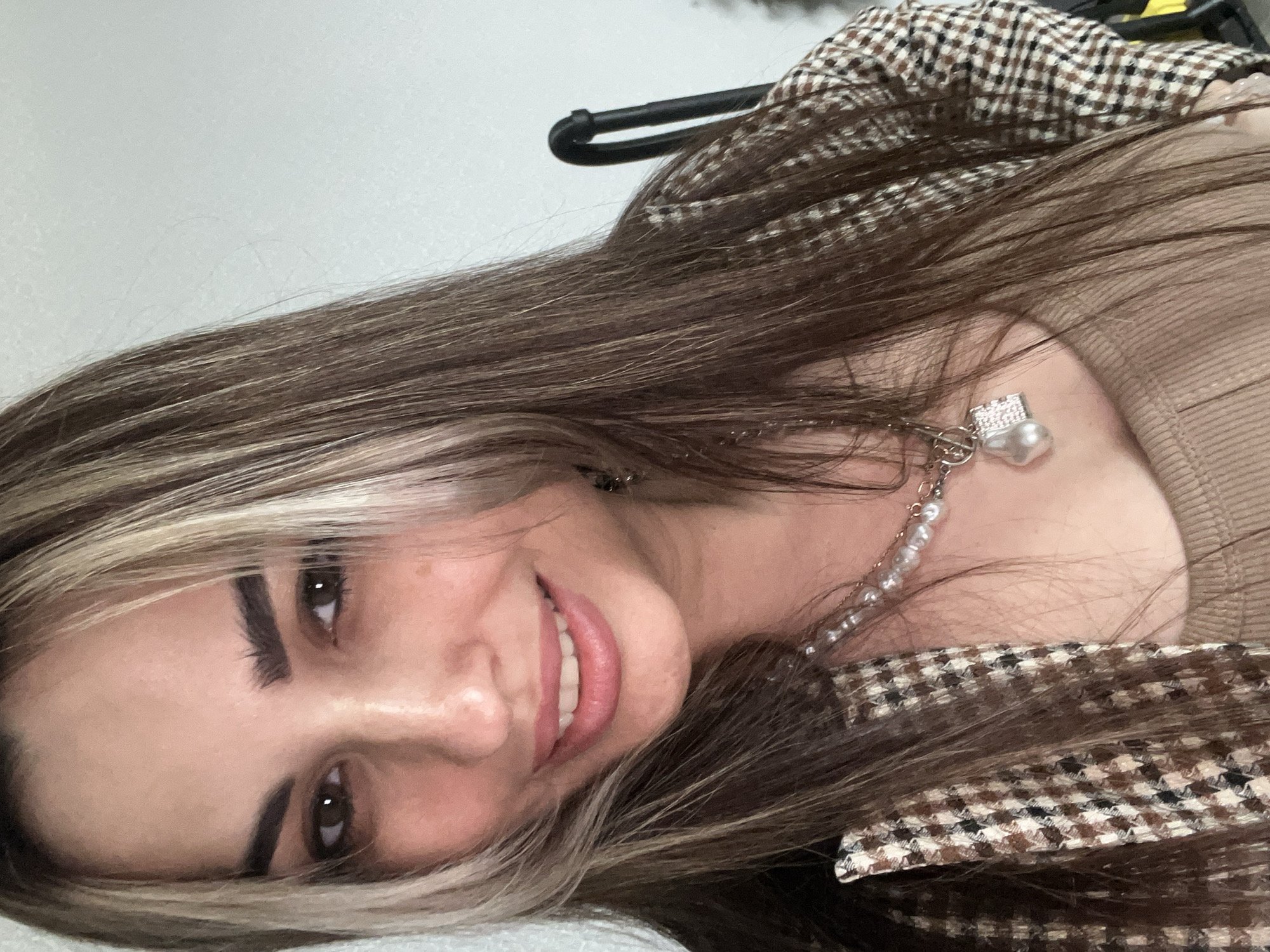 A close-up, chest-up portrait features a smiling young adult woman with fair skin, dark brown eyes, and prominent dark eyebrows. Her long, straight hair is predominantly brown with distinct light blonde streaks framing her face and interspersed throughout. She smiles directly at the viewer, revealing her teeth, and wears light makeup accentuating her eyes and lips.
She is dressed in a brown, black, and off-white houndstooth-patterned blazer, worn over a plain, ribbed, light brown top. A layered necklace adorns her neck, featuring a strand of small white pearls and a silver-toned chain with a toggle clasp. This chain supports two pendants: a small, rectangular, possibly pavé-set charm, and a larger, irregularly shaped white baroque pearl.
The background is a plain, light-colored wall, indicative of an indoor setting. A dark, curved metal object, possibly a furniture arm or leg, is visible on the far right. The location is an unknown indoor setting.