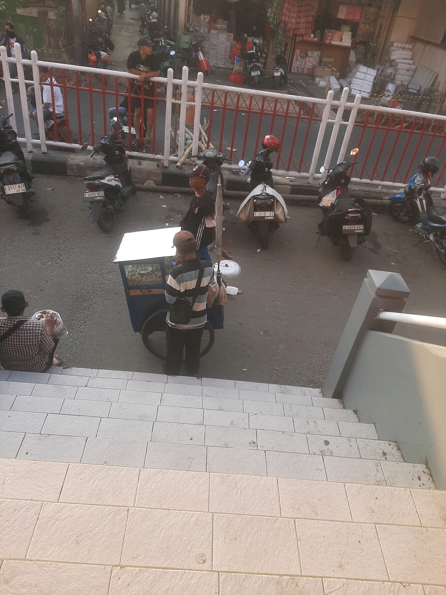 In the city of Bekasi, Indonesia, an outdoor scene unfolds with a street lined by a red and white fence. The foreground features a set of stairs leading down from a building, with two distinct types of tiling visible. On the street, several motorcycles are parked.
A man in a plaid shirt sits on the steps of the stairs, holding a plastic bag filled with food. He is facing away from the camera and towards a street food cart. The cart is blue and white, with a glass display case showing food items. A vendor, wearing a striped shirt and a cap, stands behind the cart, attending to his goods.
Behind the vendor and the cart, several other individuals are visible, some standing near motorcycles. One person is wearing a black shirt and shorts, leaning on the fence. Another person in the background wears a red helmet. The environment suggests a busy urban setting, likely during daytime given the lighting. The street itself appears to be asphalt.