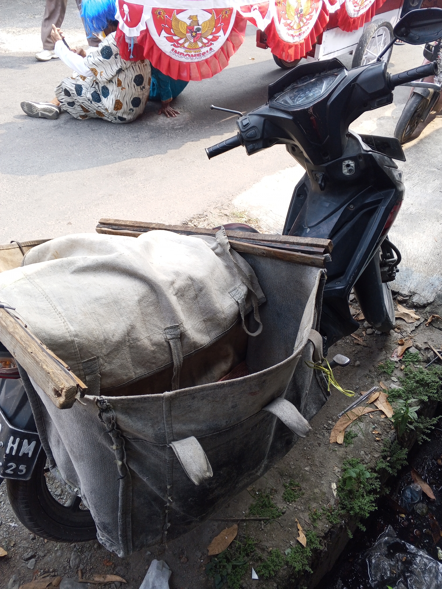 In the city of Ciruas, Indonesia, on a paved street, a black scooter is parked. It has a large canvas bag attached to the rear, designed for carrying goods. The bag is filled with a white tarp, which is partially open. The scooter's handlebars and dashboard are visible. To the left of the scooter, a person in a blue wig and a patterned dress is sitting on the ground. They are looking at a mobile phone in their hands. Behind this person is a cart draped with red and white banners, one of which features the Indonesian national emblem, the Garuda Pancasila. Further in the background, another person's legs are visible. The ground around the scooter and the cart is a mix of dirt and asphalt. There are some dry leaves and small plants growing on the side of the road. A small trench filled with dark water is also present to the right.