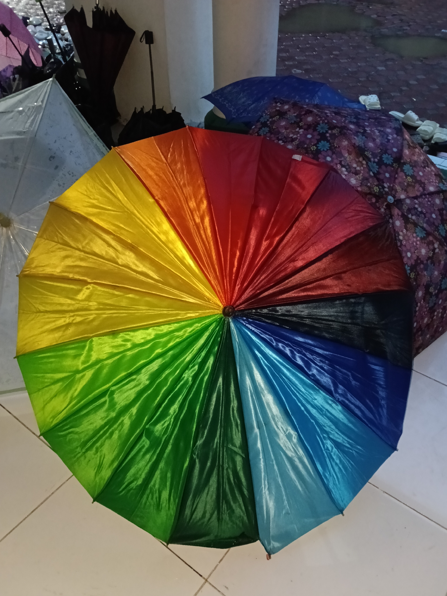 A colorful umbrella dominates the foreground on a light-colored tiled surface. The umbrella is composed of sections in a rainbow pattern of colors from yellow to blue. Behind the rainbow umbrella are other umbrellas in varying states of closure and color. A clear umbrella and a brown umbrella are to the left of the rainbow umbrella. A blue and a patterned umbrella are to the right. The location appears to be indoors near a doorway, as indicated by the glimpse of a wet brick surface visible in the upper right. The image was taken in Lhokseumawe, Indonesia.