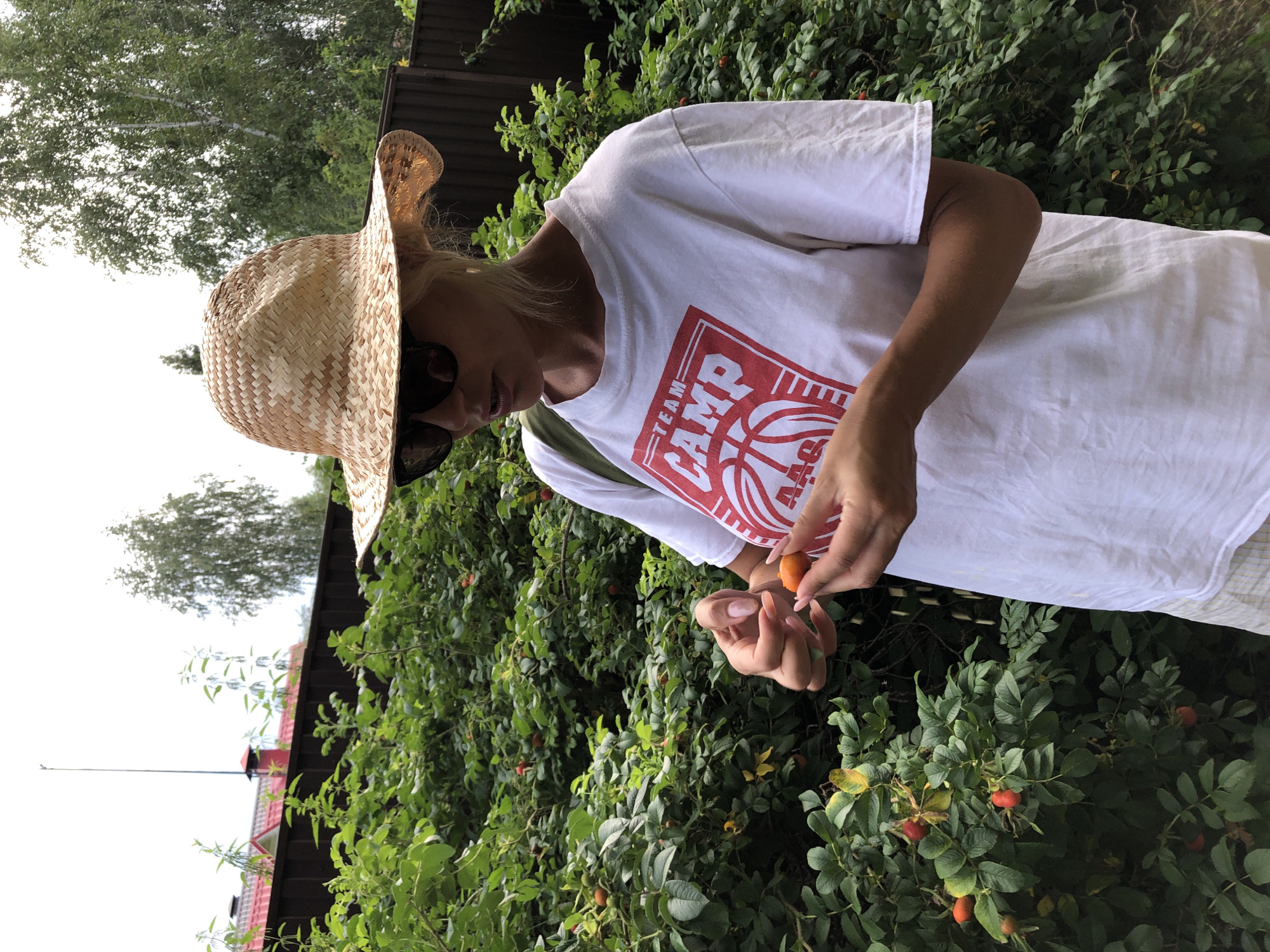 A person wearing a straw hat and sunglasses is standing outdoors in a garden, holding a small orange berry in their hands. They are wearing a white t-shirt with a red graphic on it, and a portion of a checkered skirt is visible. Behind them is dense green foliage with several red berries on the bushes. A wooden fence and trees are visible in the background. The scene appears to be in the city of Садко Снт (макеиха), Russia.