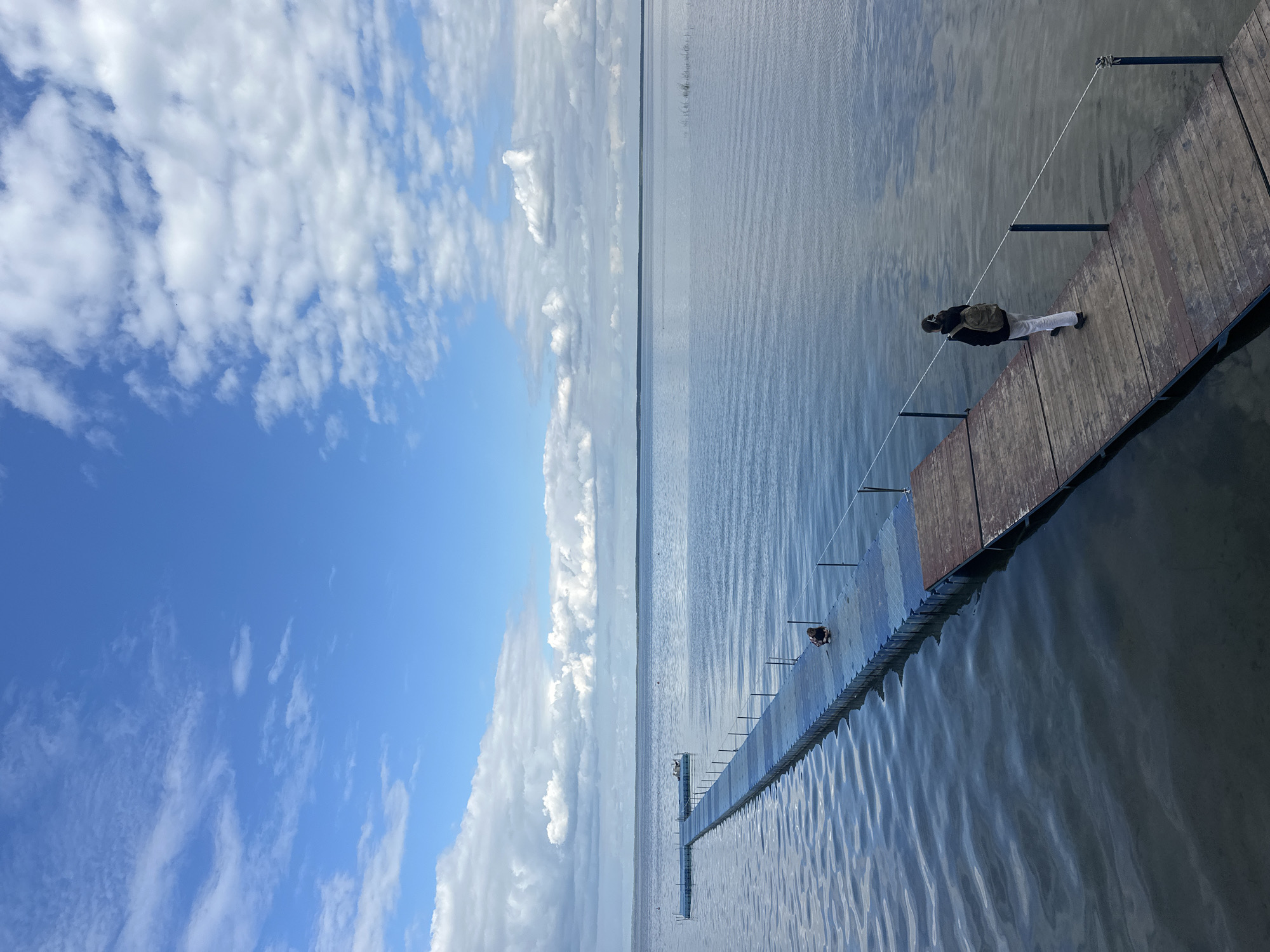 The image shows a long, narrow pier extending into a wide, calm body of water under a bright blue sky with scattered white clouds. A person wearing a backpack is walking away from the viewer on the pier. The pier is constructed of wooden planks and sections of blue plastic modules. Further out on the water, there is a smaller, floating platform. The horizon is visible in the distance with a faint landmass. The water is slightly rippled, reflecting the sky and clouds. The scene appears to be in a natural, open-air environment, likely a lake or large river. The specific location is in the Нарацкі сельскі Савет, Belarus, suggesting a rural or park-like setting associated with Lake Naroch.