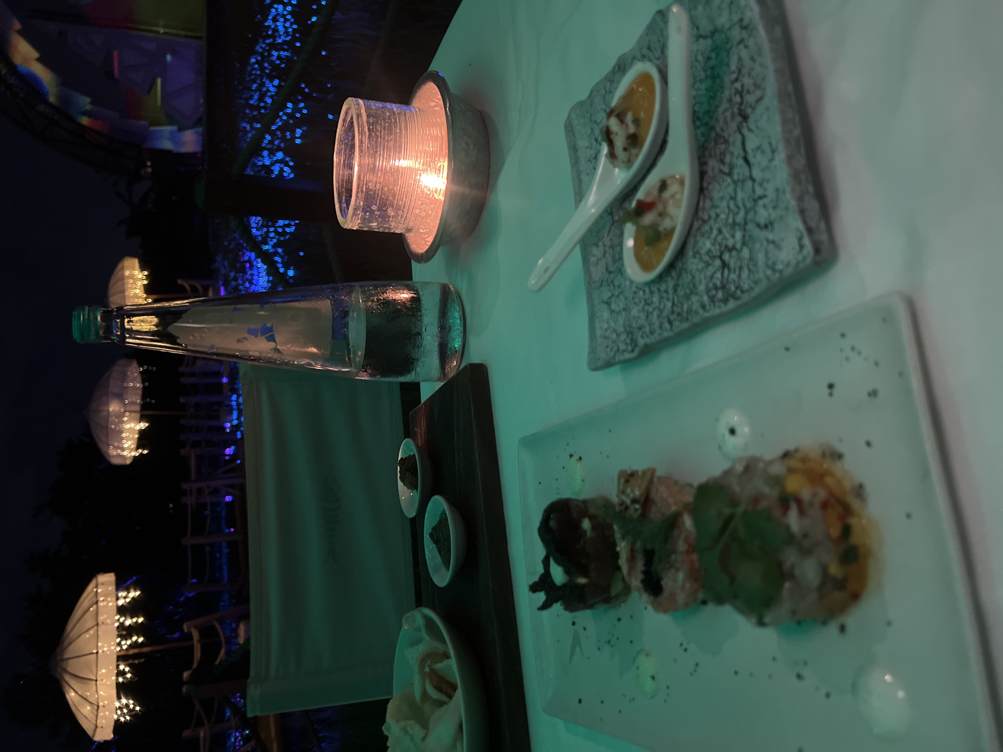 This image captures a nighttime outdoor dining scene, likely at an upscale restaurant or resort in North Kuta, Indonesia. The setting is characterized by a cool, ambient blue-green light, contrasting with the warm, orange glow emanating from a candle in a clear glass holder on the table.
The foreground focuses on a well-appointed table covered with a light-colored cloth. Prominently featured is a tall, slender glass bottle of water with a green cap. Various culinary items are presented: a rectangular grey plate holds two white ceramic spoons, each containing an appetizer featuring a yellow sauce and diced ingredients. Adjacent to this, a white rectangular plate displays several artfully arranged portions of food, possibly sushi or modern tapas, garnished with herbs and dark speckles of spices. A wooden serving board holds two small white bowls with dark contents, presumably sauces or condiments, and part of another white bowl containing bread or crackers is visible.
The background reveals a dark night sky illuminated by numerous small blue lights, which could be decorative string lights or lighting from water features. Standing out are several large, distinctive white structures, resembling illuminated umbrella-like lanterns or jellyfish, casting a warm yellow light. These structures contribute significantly to the exotic and atmospheric ambiance of the outdoor space. The scene suggests a serene and elegant dining experience. No legible text is visible in the image.