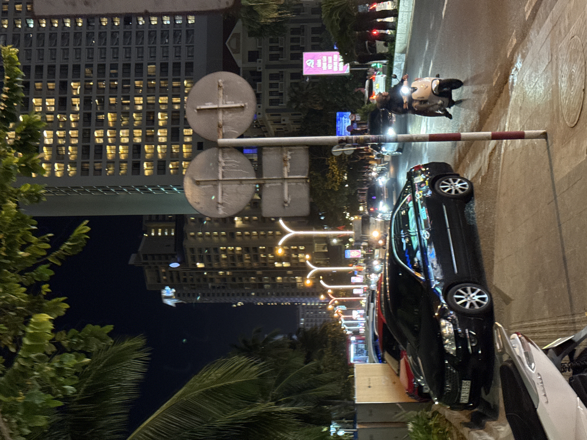 The image depicts a nighttime street scene in Nha Trang, Vietnam, likely along a coastal boulevard. Towering, brightly lit buildings, characteristic of a resort city, dominate the background. In the foreground, a black sedan is parked on the side of the road. A person is riding a scooter, heading away from the camera. Streetlights illuminate the road, reflecting on its wet surface, suggesting recent rain. Palm trees and foliage frame the scene, common in tropical coastal environments like Nha Trang. The overall impression is a lively urban nightscape.